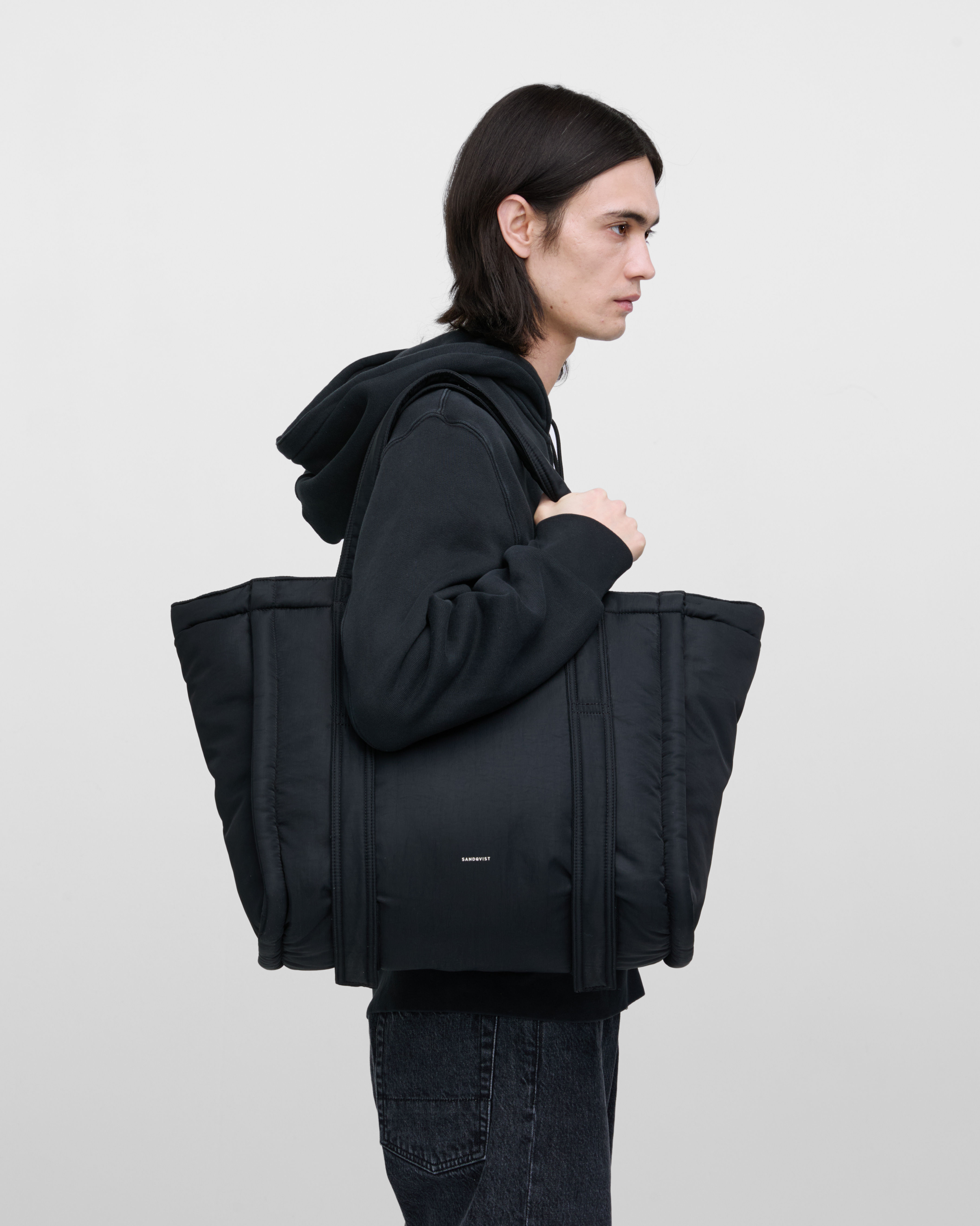Male model wearing Sandqvist Curve Tote Bag, black. Side view, wearing the bag on one shoulder.