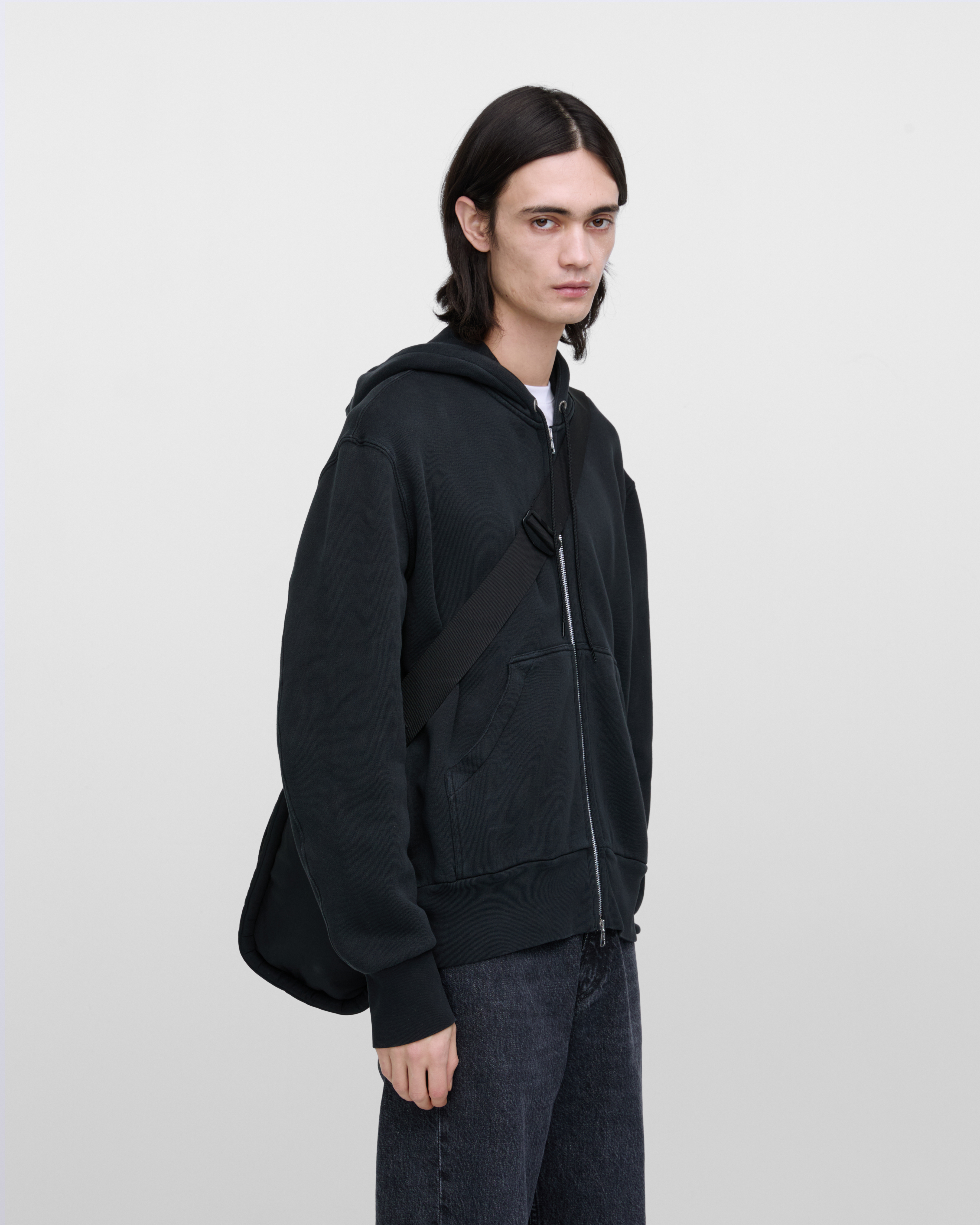 Male model wearing Sandqvist Curve Tote Bag, black. Front view, wearing the bag on one shoulder crossbody, with the bag in the back.