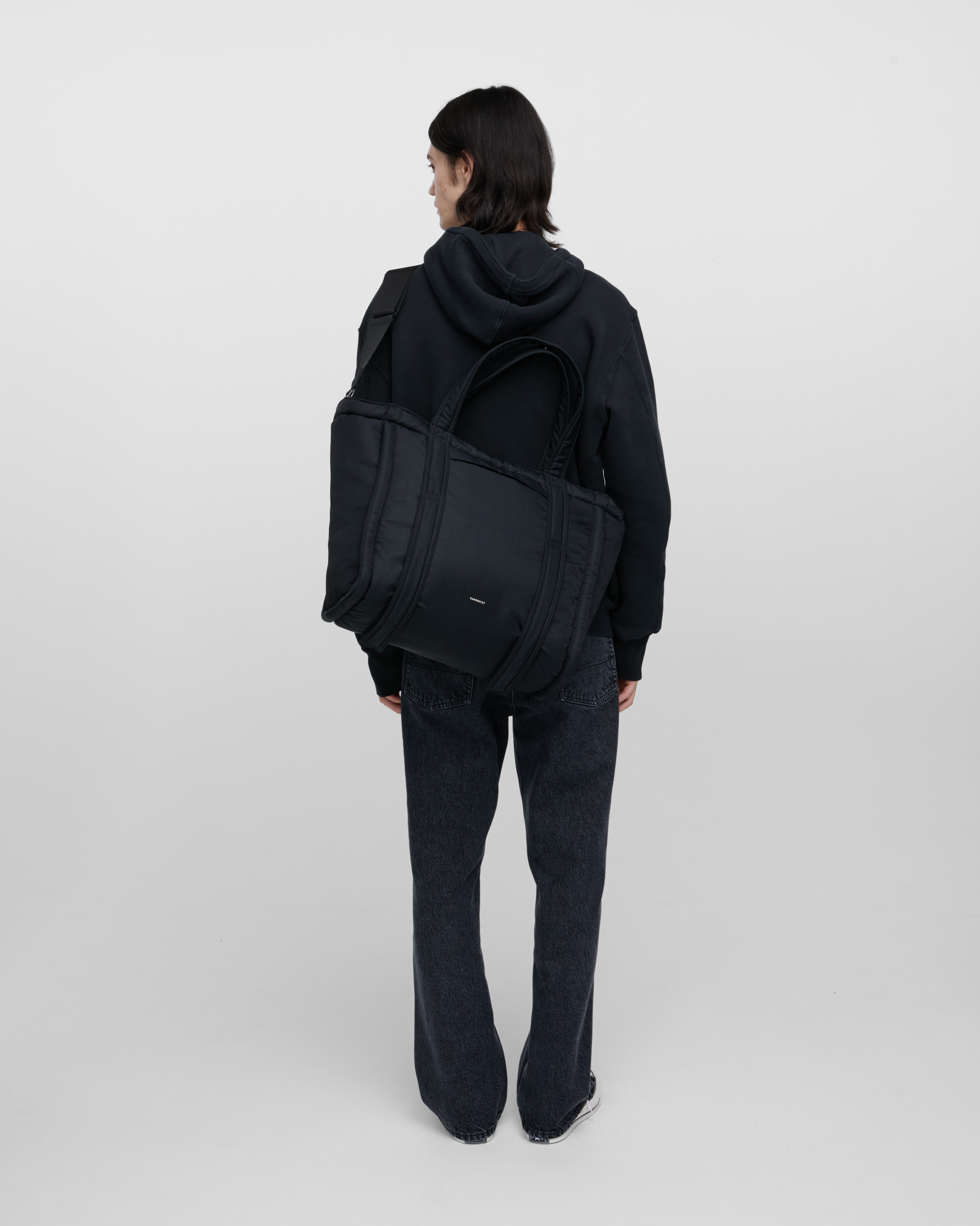 Male model wearing Sandqvist Curve Tote Bag, black. Back view, wearing the bag on one shoulder crossbody, with the bag in the back.
