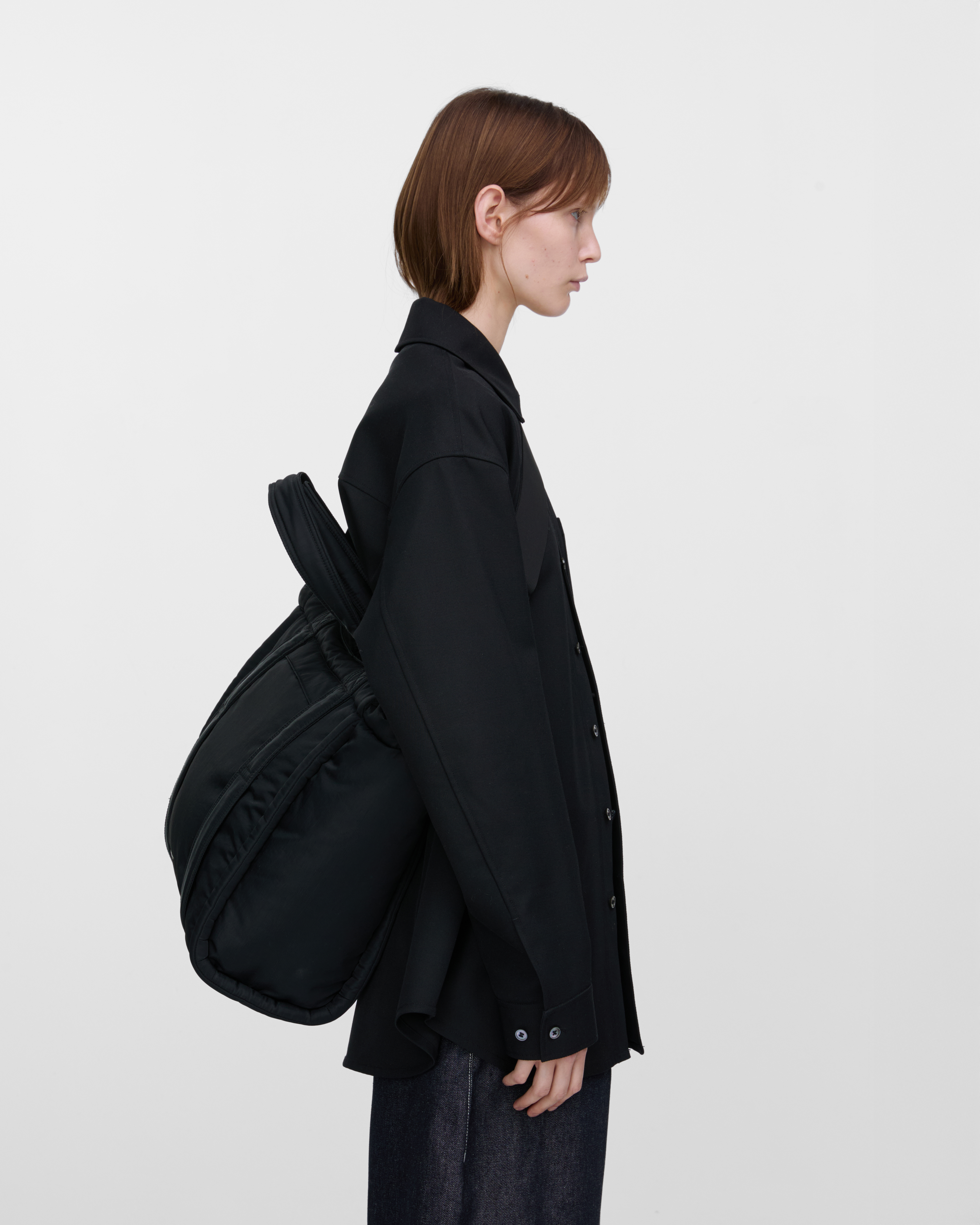 Female model wearing Sandqvist Curve Tote Bag, black. Side view, wearing the bag on one shoulder crossbody, with the bag in the back.