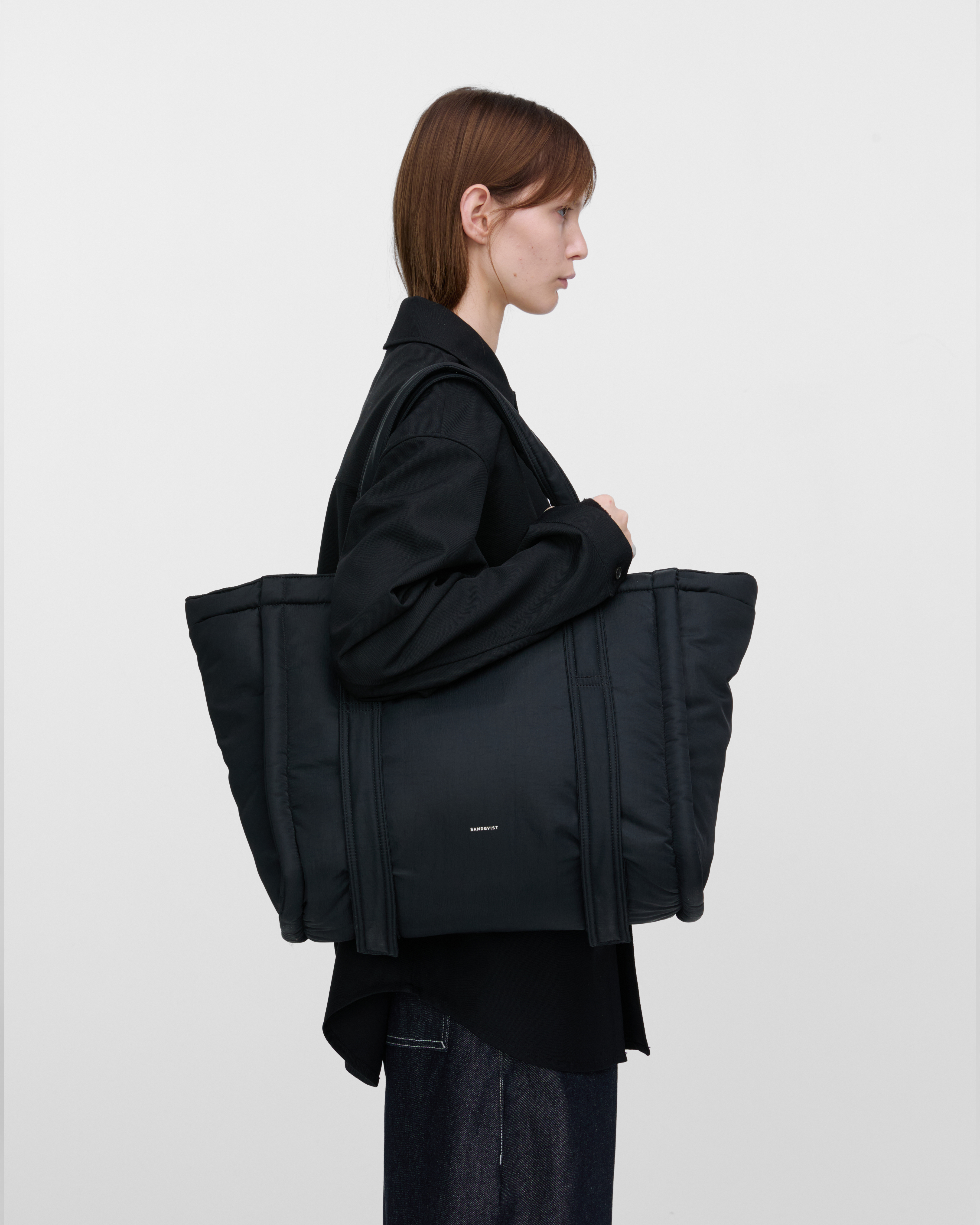 Female model wearing Sandqvist Curve Tote Bag, black. Side view, wearing the bag on one shoulder.