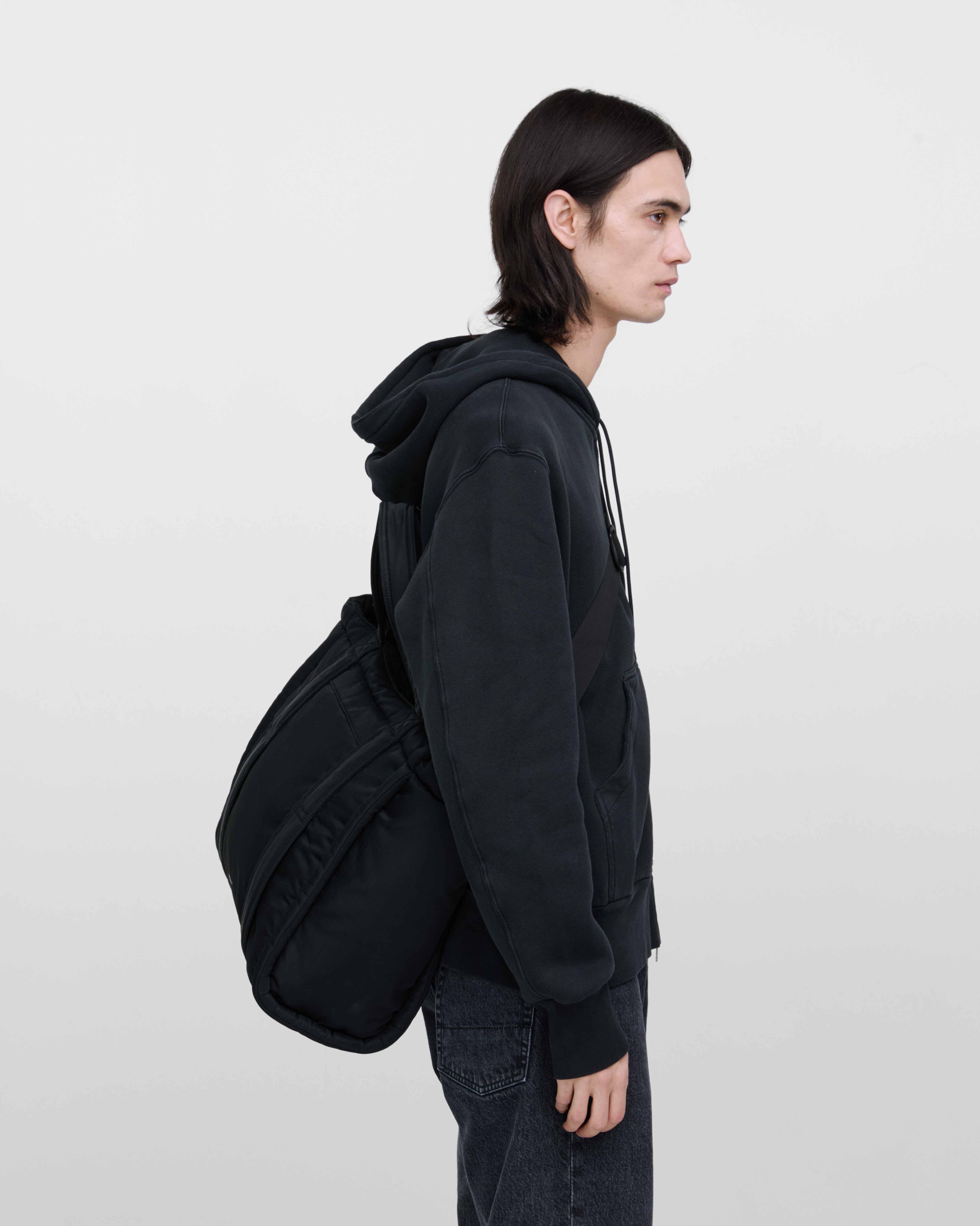 Male model wearing Sandqvist Curve Tote Bag, black. Side view, wearing the bag on one shoulder crossbody, with the bag in the back.
