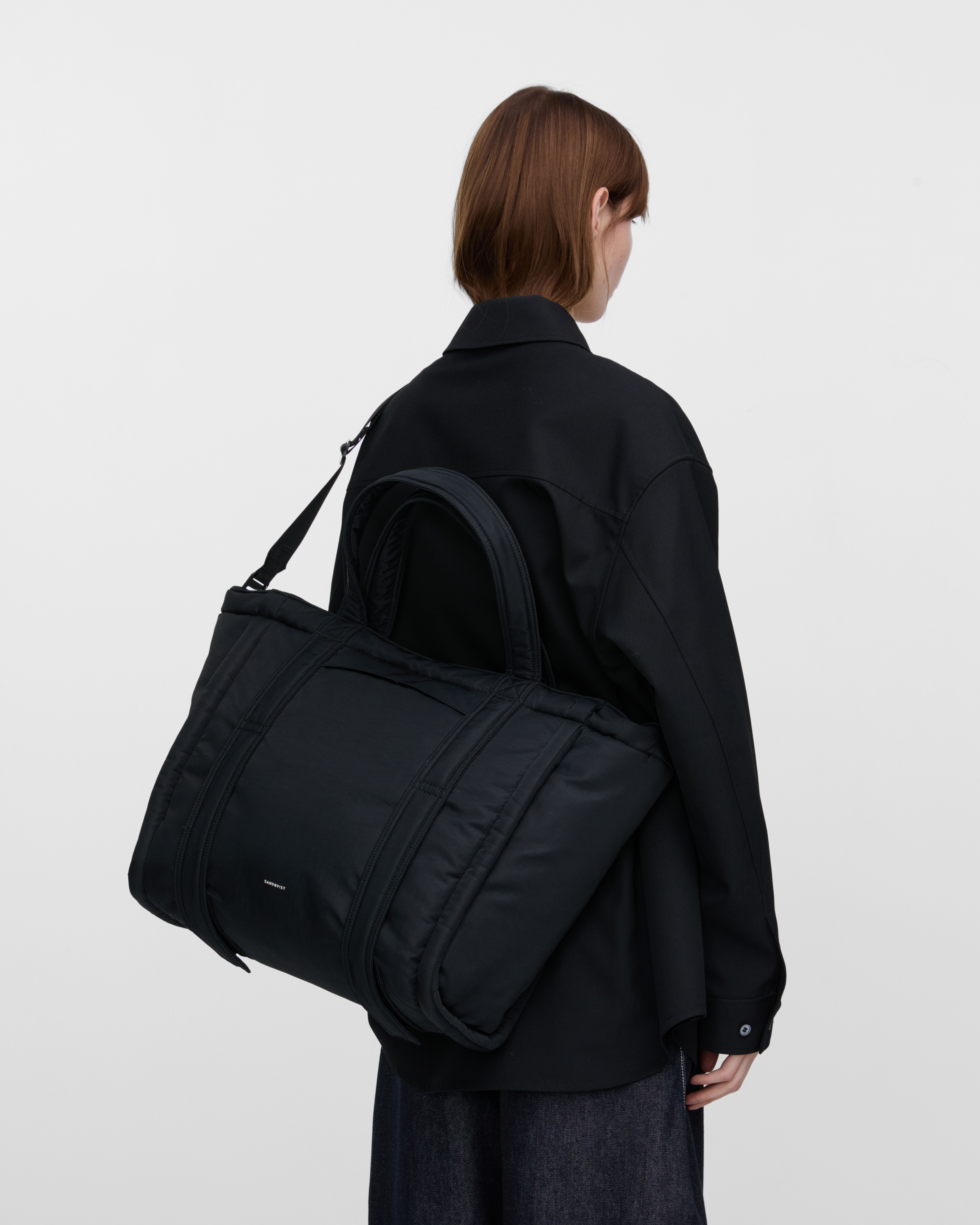 Female model wearing Sandqvist Curve Tote Bag, black. Back view, wearing the bag on one shoulder crossbody, with the bag in the back.