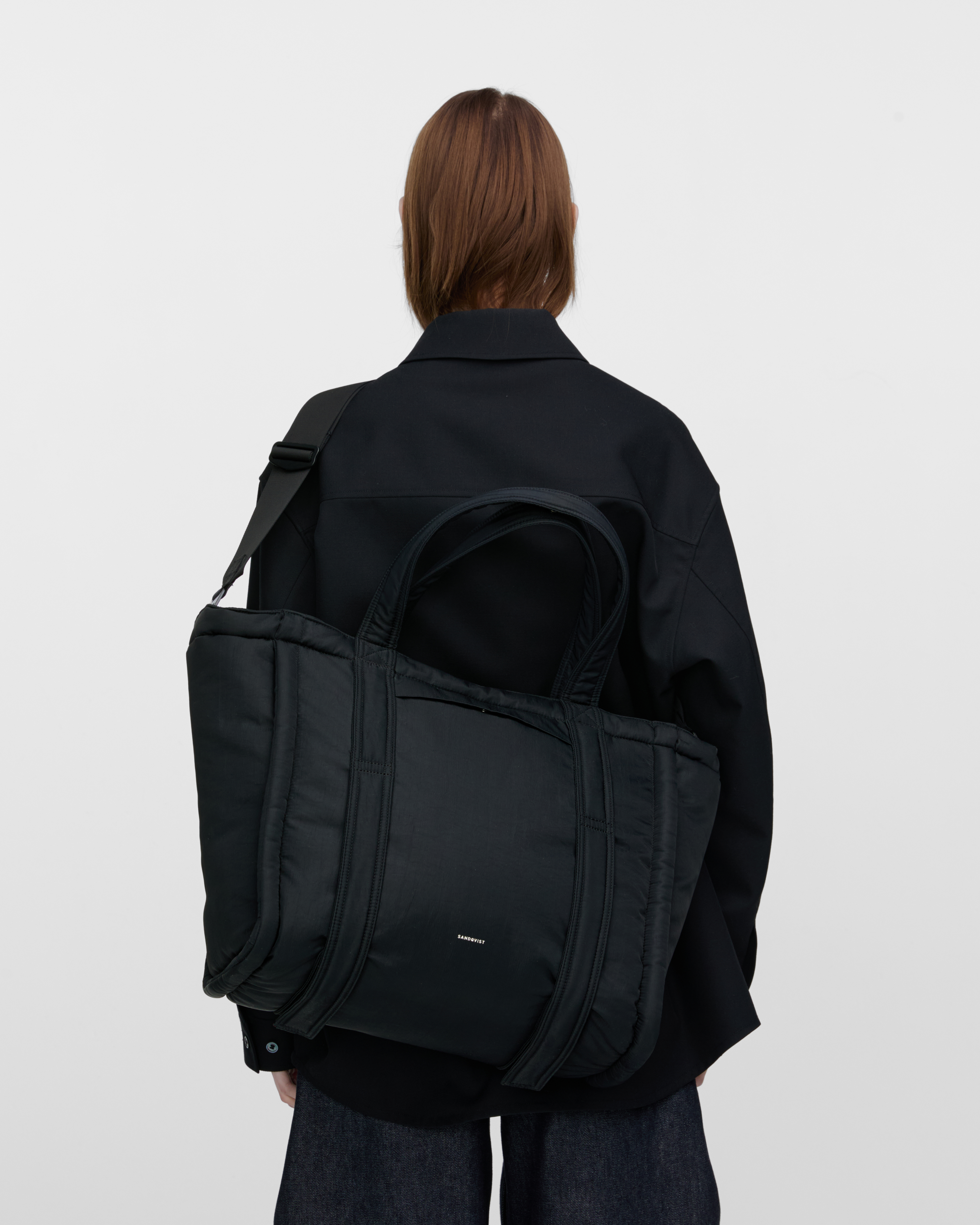 Female model wearing Sandqvist Curve Tote Bag, black. Back view, wearing the bag on one shoulder crossbody, with the bag in the back.