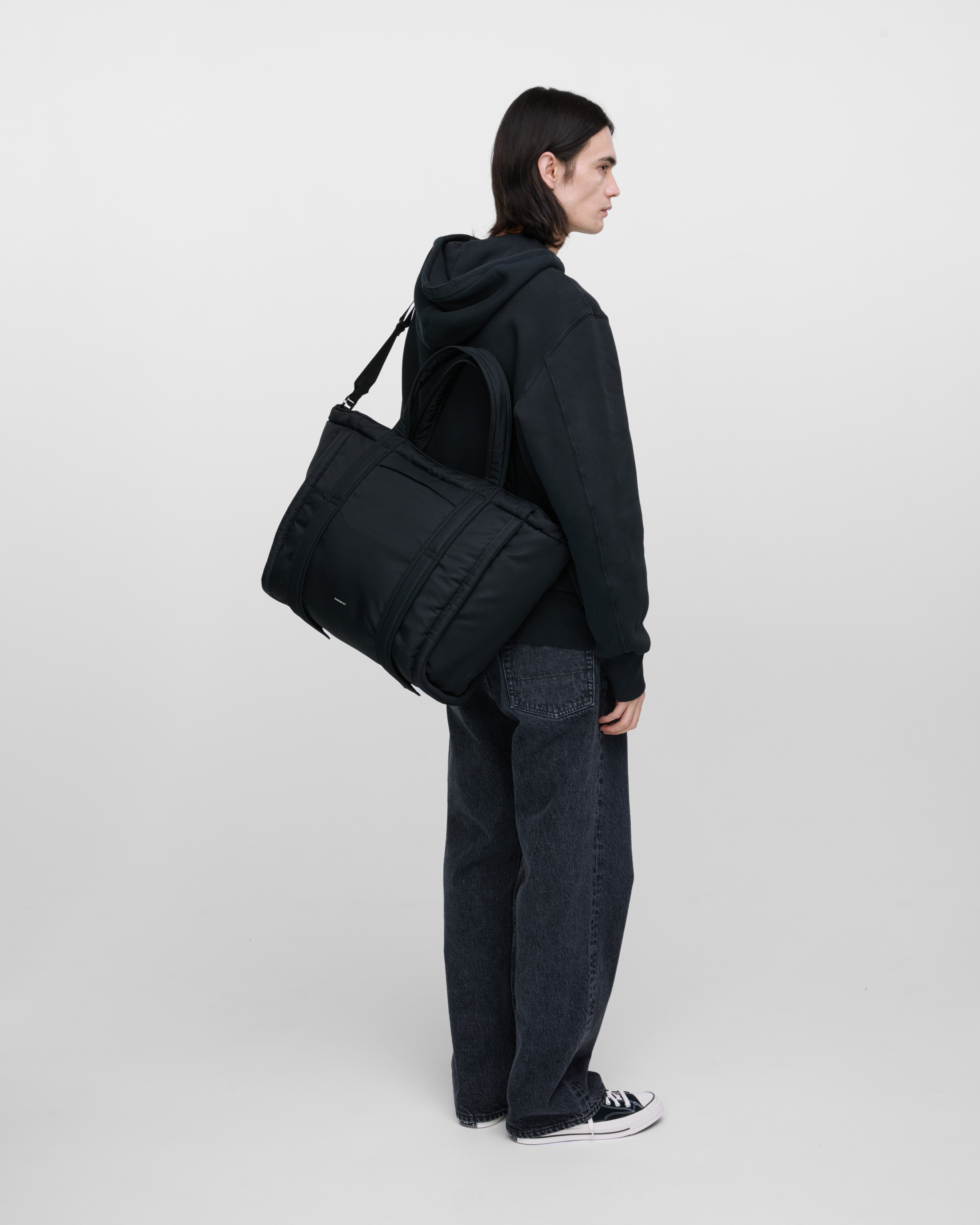 Male model wearing Sandqvist Curve Tote Bag, black. Back view, wearing the bag on one shoulder crossbody, with the bag in the back.