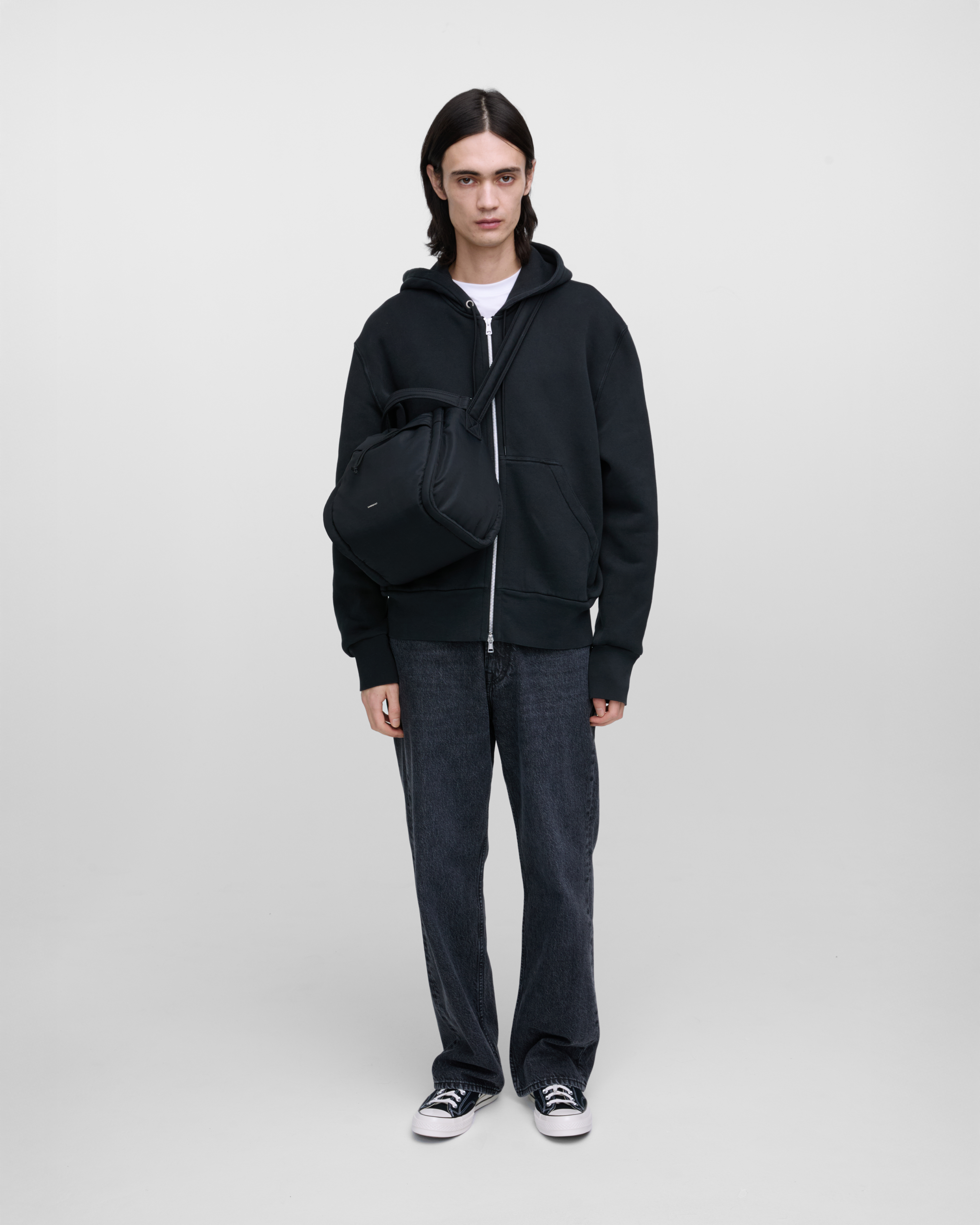 Male model wearing Sandqvist Curve Bucket Bag, black. Front view, carrying the bag on one shoulder crossbody with the bag in the front.