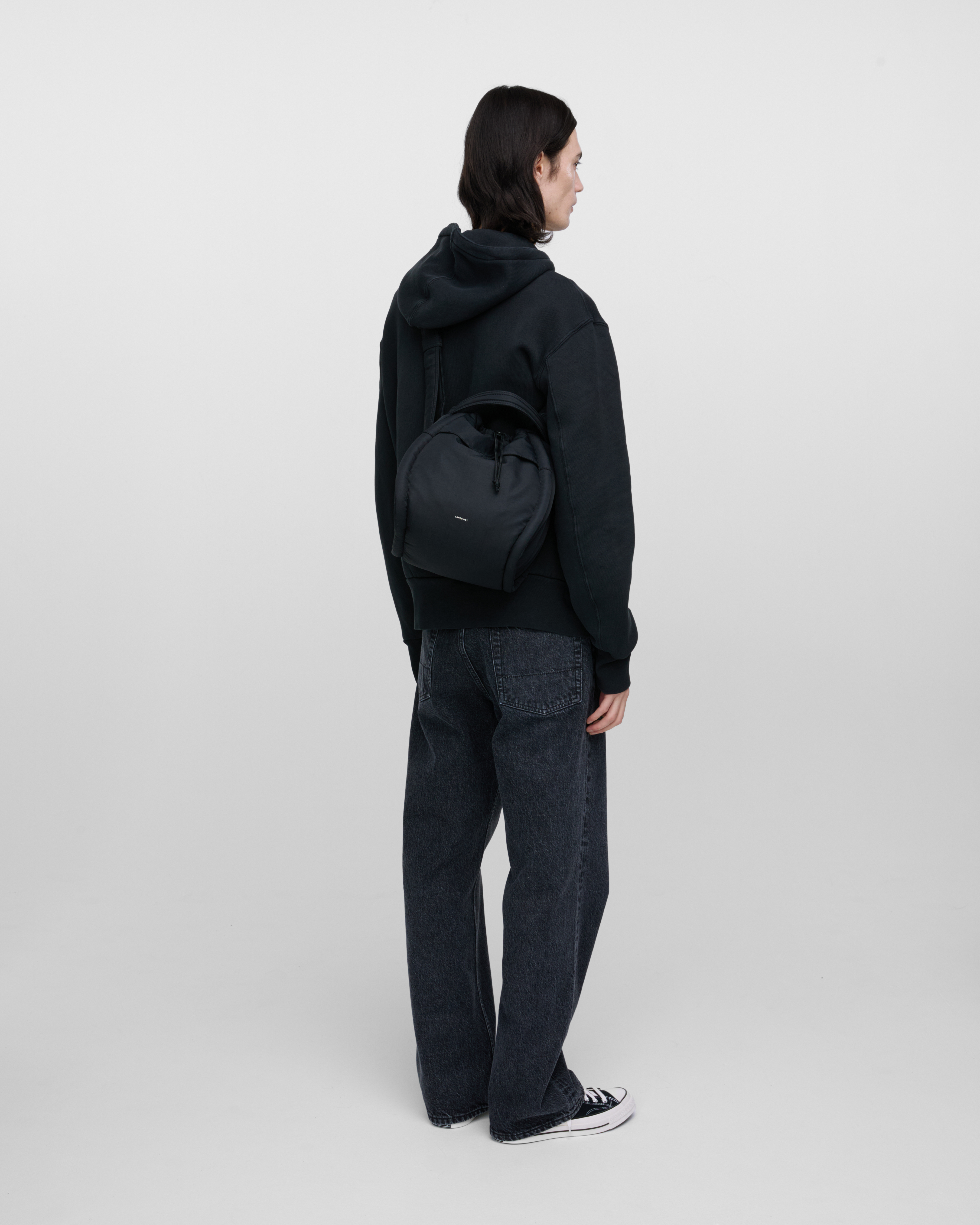 Male model wearing Sandqvist Curve Bucket Bag, black. Back view, carrying the bag on one shoulder crossbody with the bag on the back.