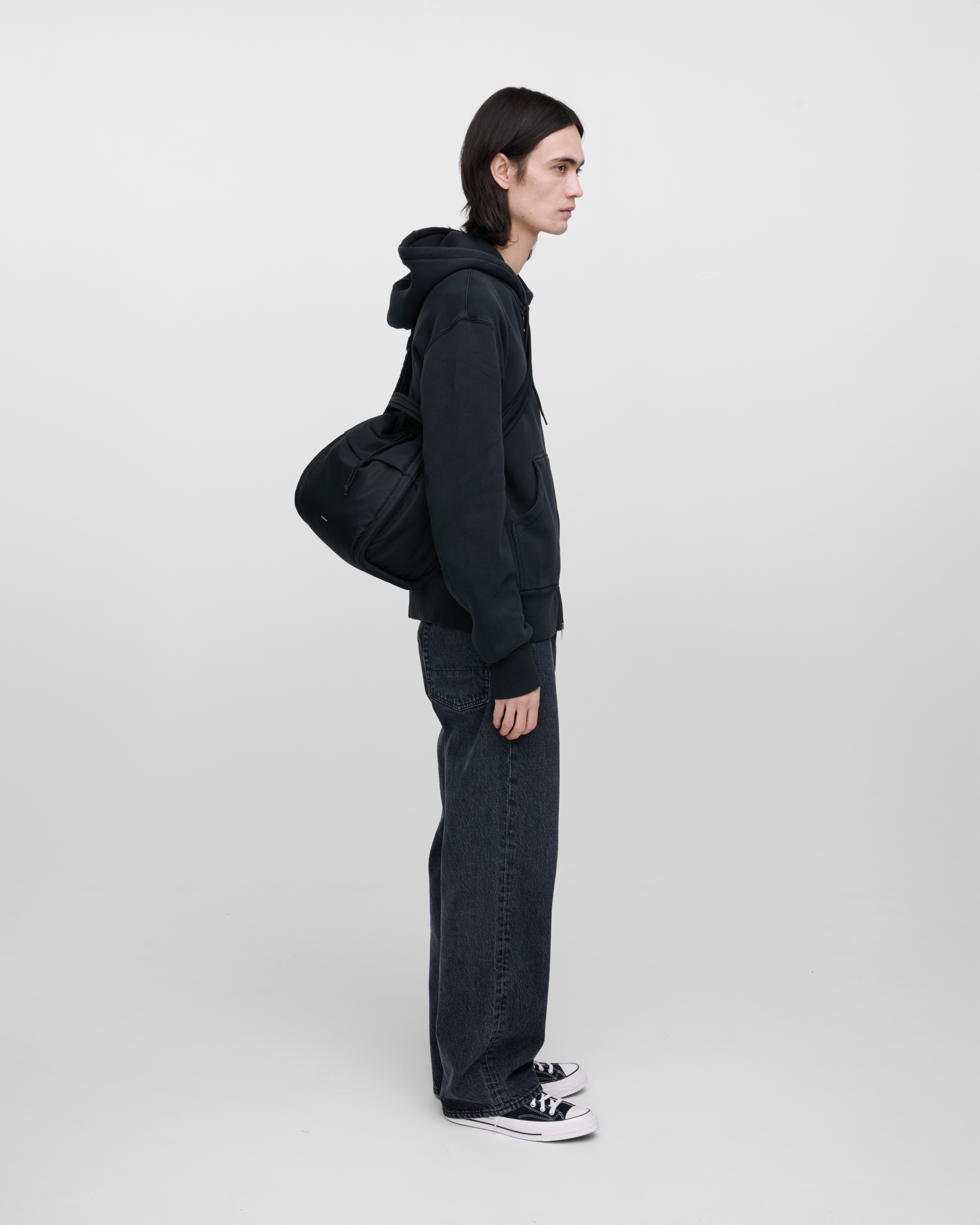 Male model wearing Sandqvist Curve Bucket Bag, black. Side view, carrying the bag on one shoulder crossbody with the bag in the back.