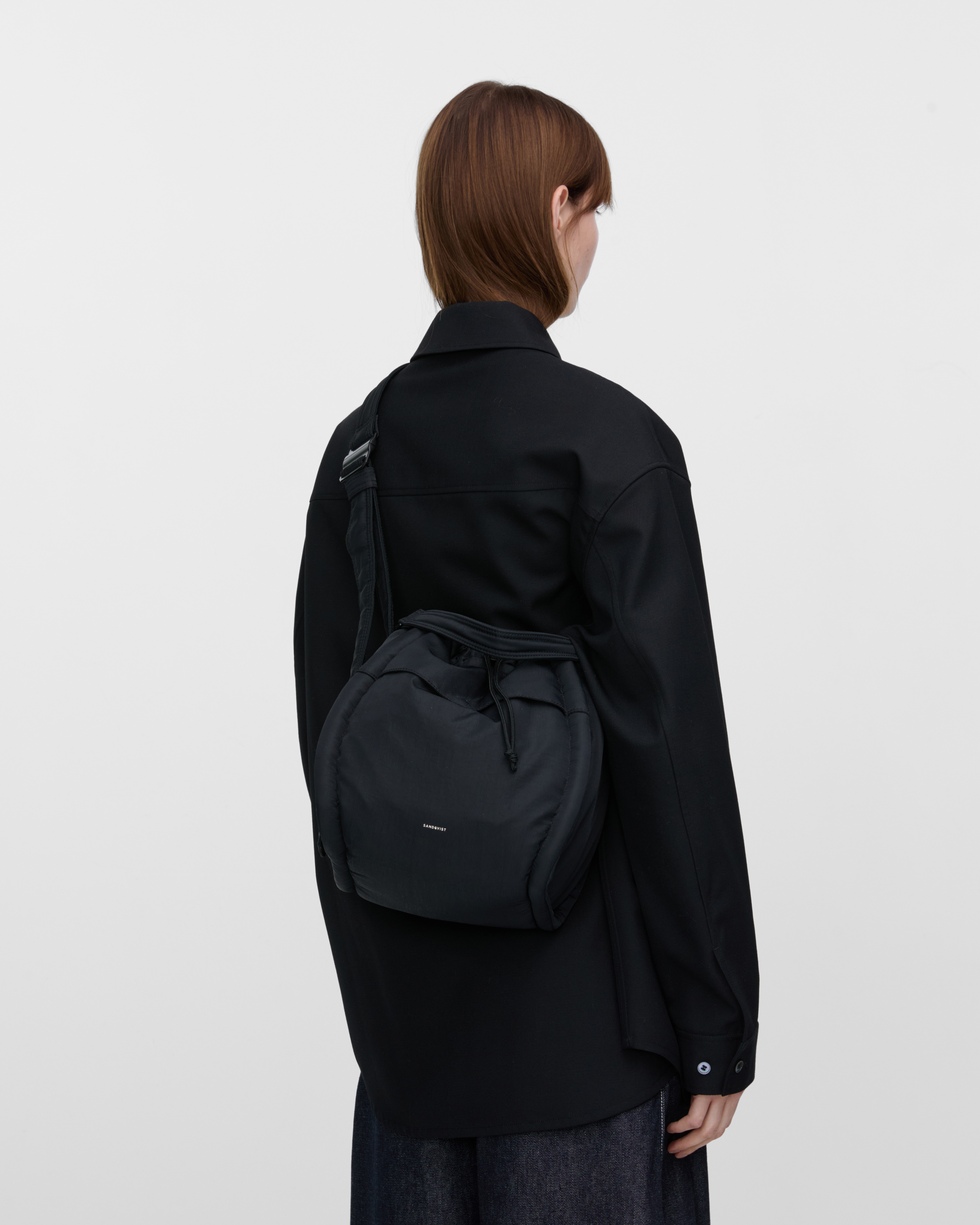 Female model wearing Sandqvist Curve Bucket Bag, black. Back view, wearing the bag crossbody with the bag on the back.