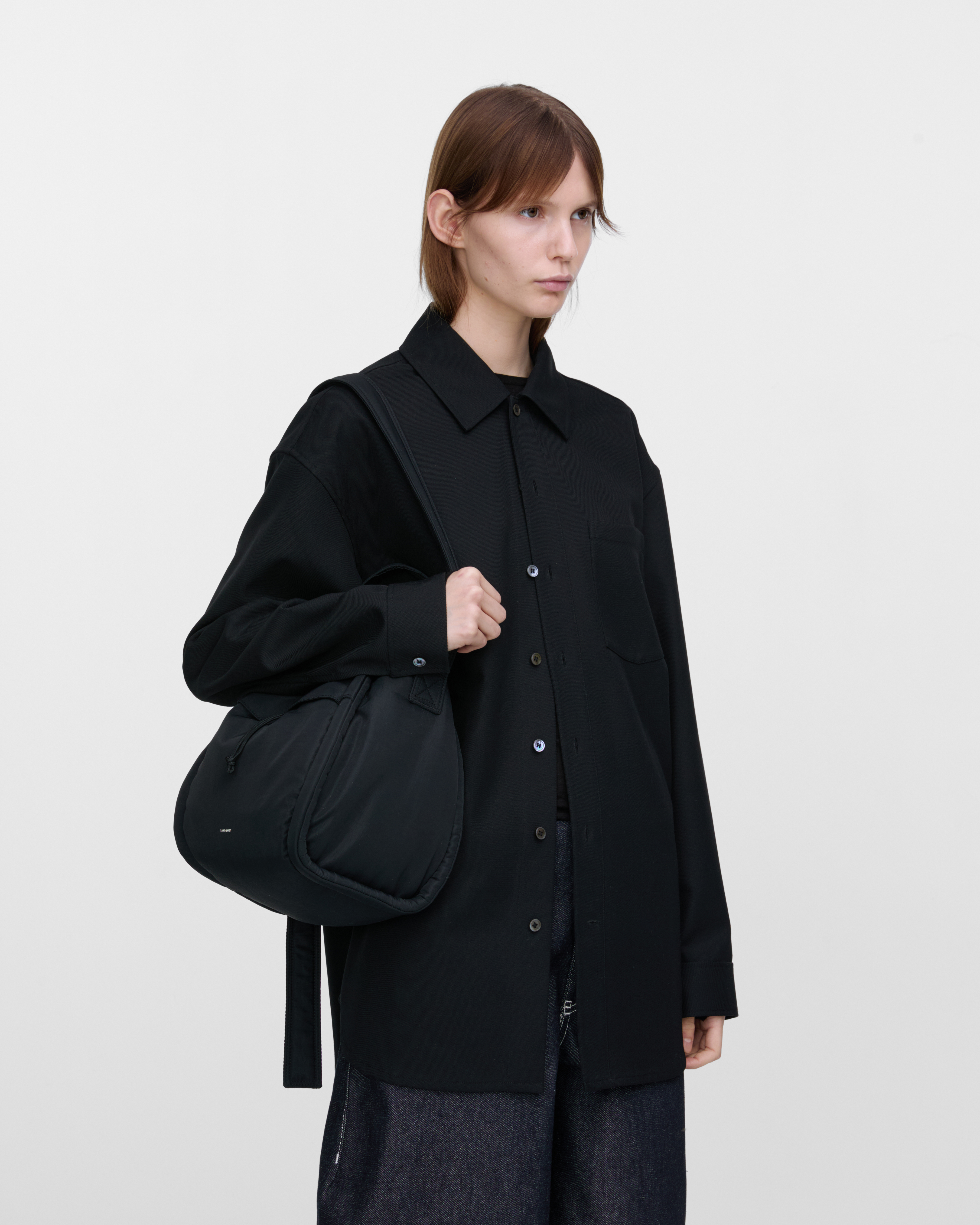 Female model wearing Sandqvist Curve Bucket Bag, black. Front view, carrying the bag on one shoulder.
