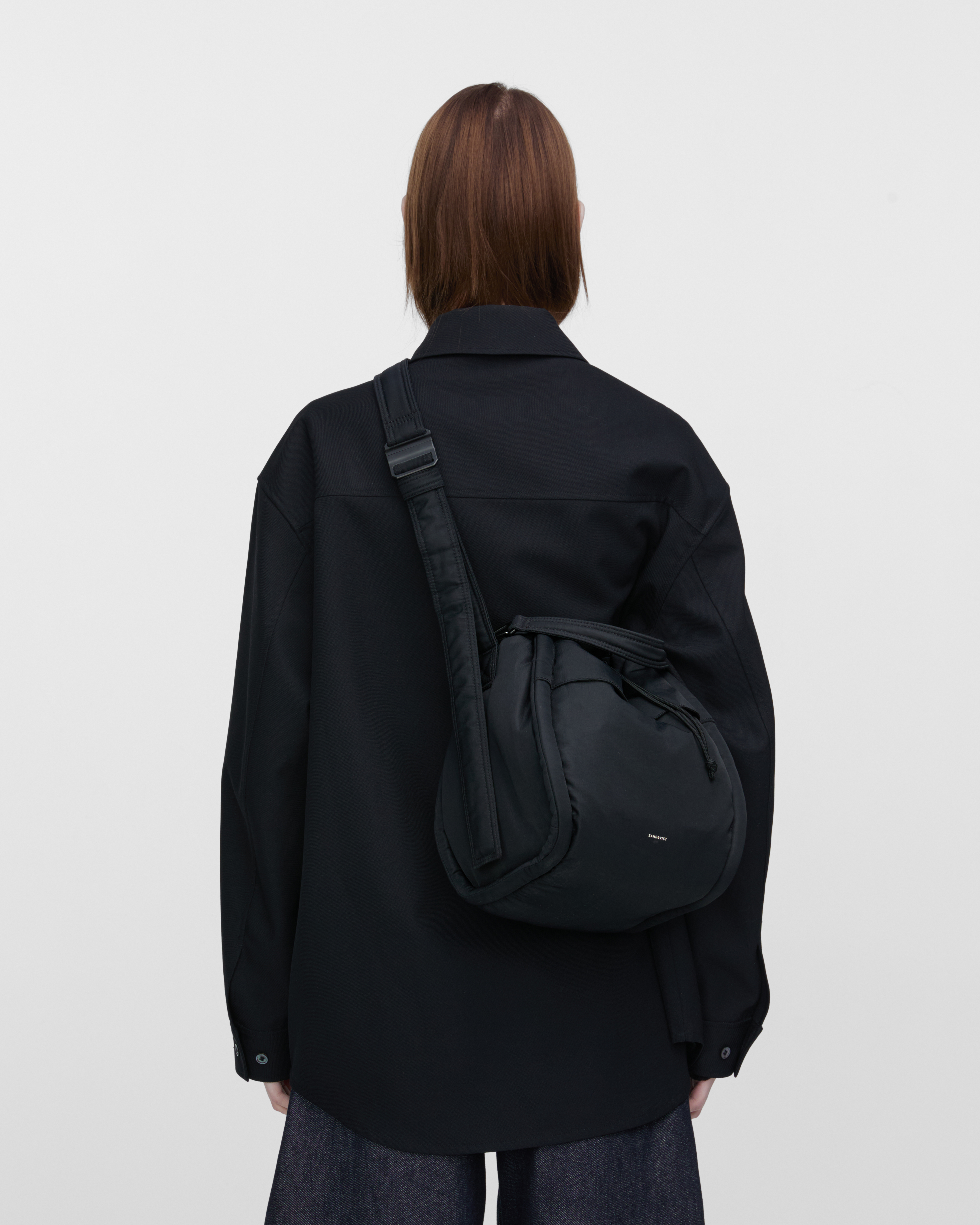 Female model wearing Sandqvist Curve Bucket Bag, black. Back view, carrying the bag crossbody with the bag on the back.