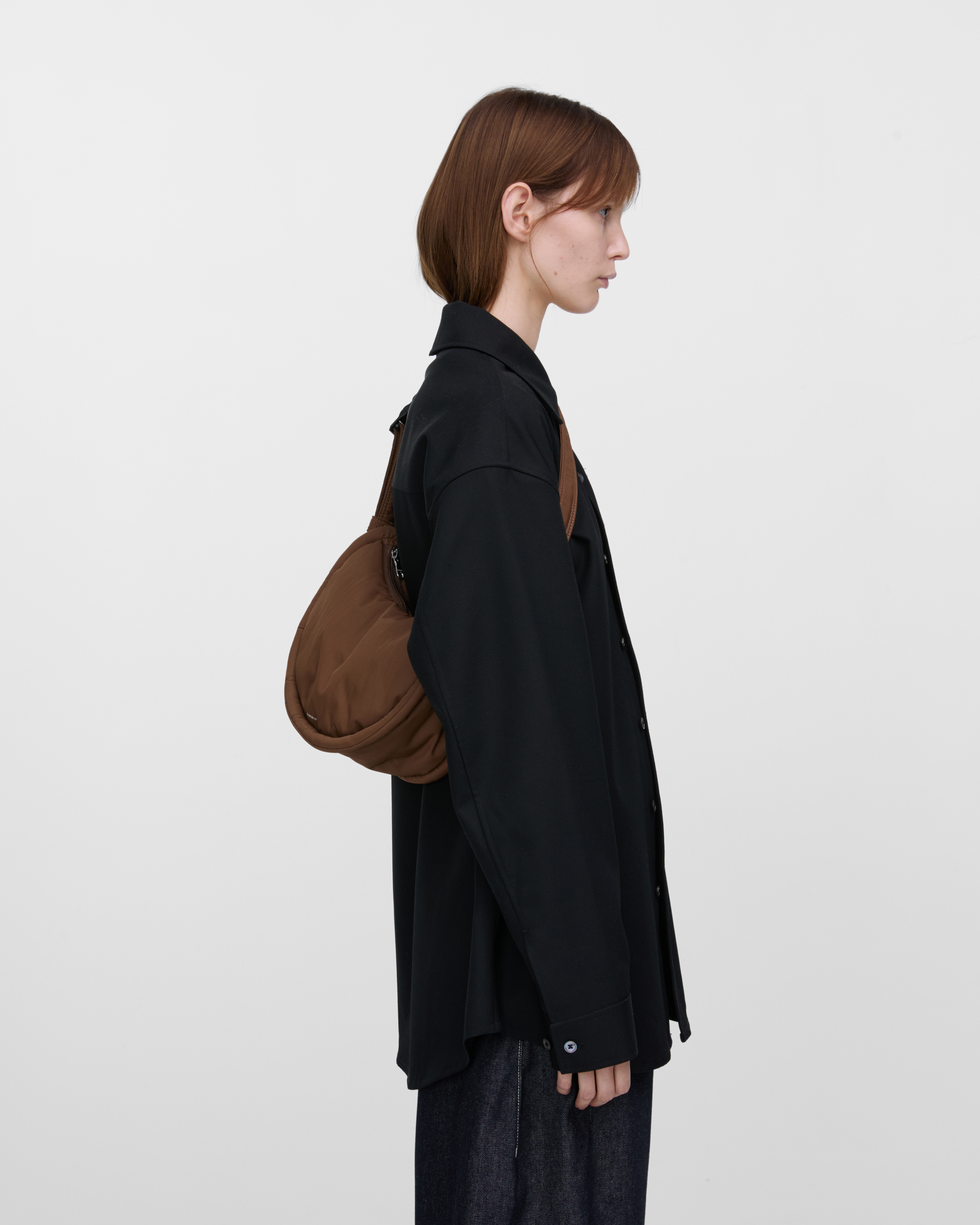 Female model wearing Sandqvist Curve Half Moon Bag Mini, brown. Side view, carrying the bag on one shoulder crossbody with the bag in the back.