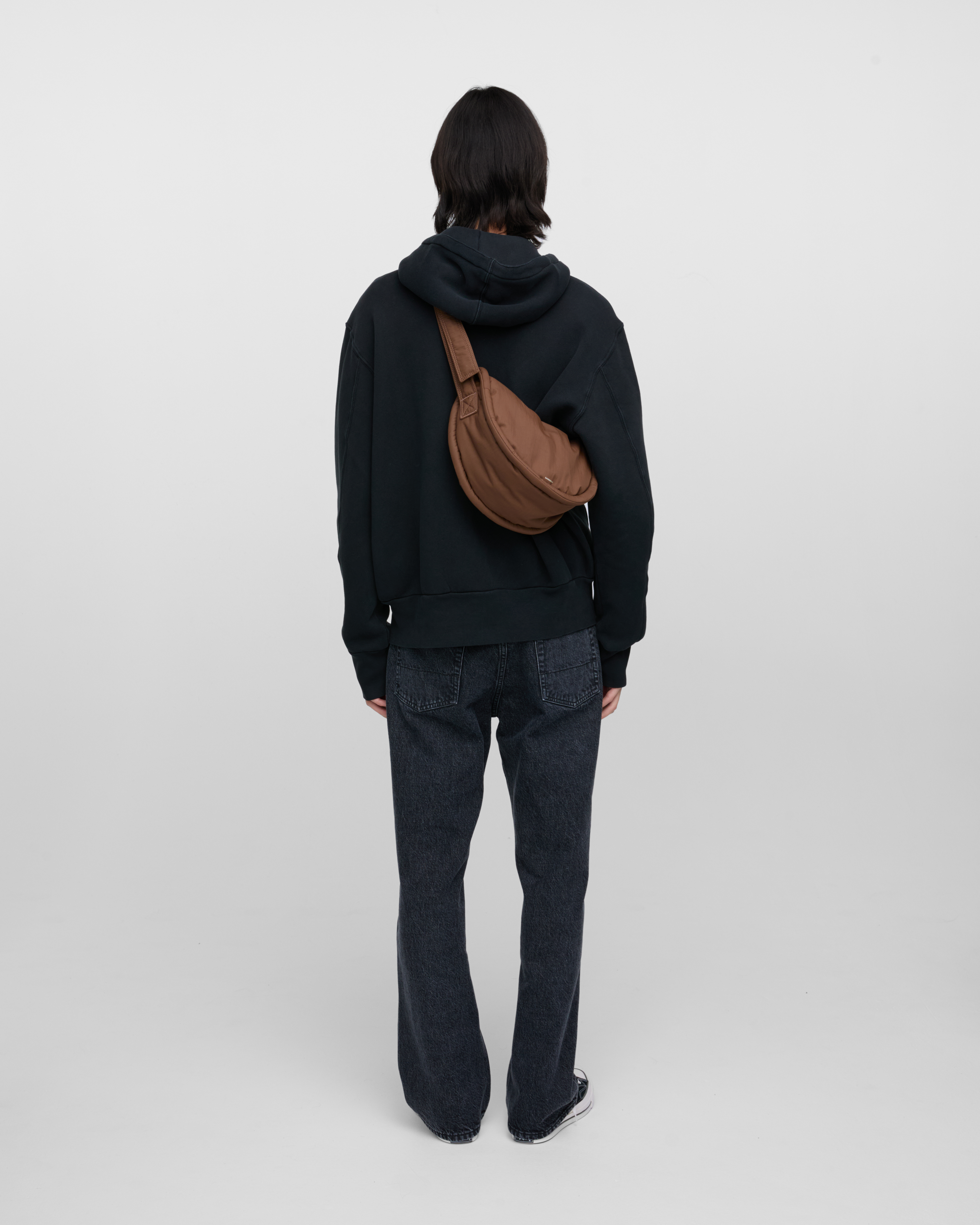 Male model wearing Sandqvist Curve Half Moon Bag Mini, brown. Back view, carrying the bag on one shoulder crossbody with the bag on the back.