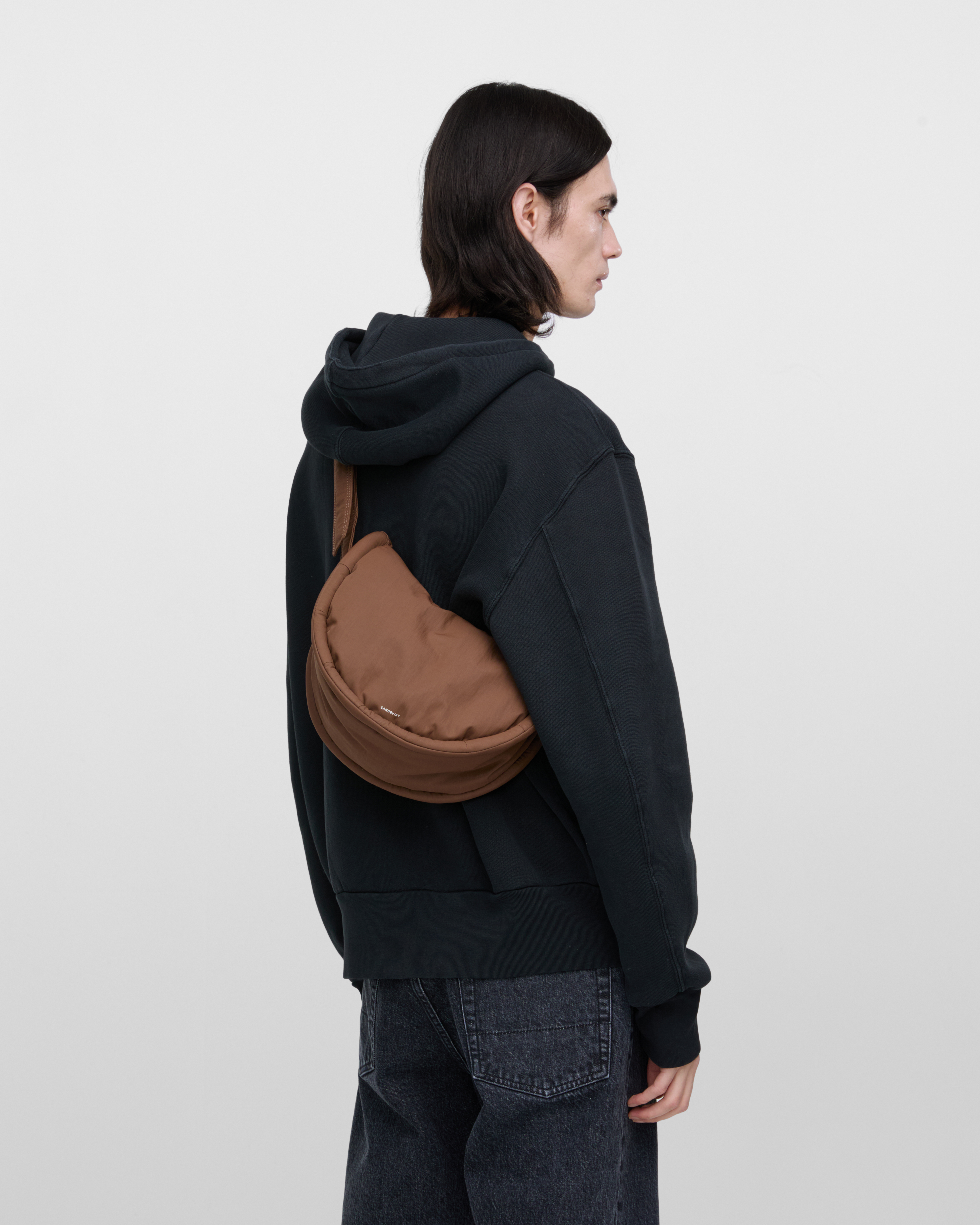 Male model carrying Sandqvist Curve Half Moon Bag Mini, brown. From behind, carrying the bag over one shoulder with the bag on his back.