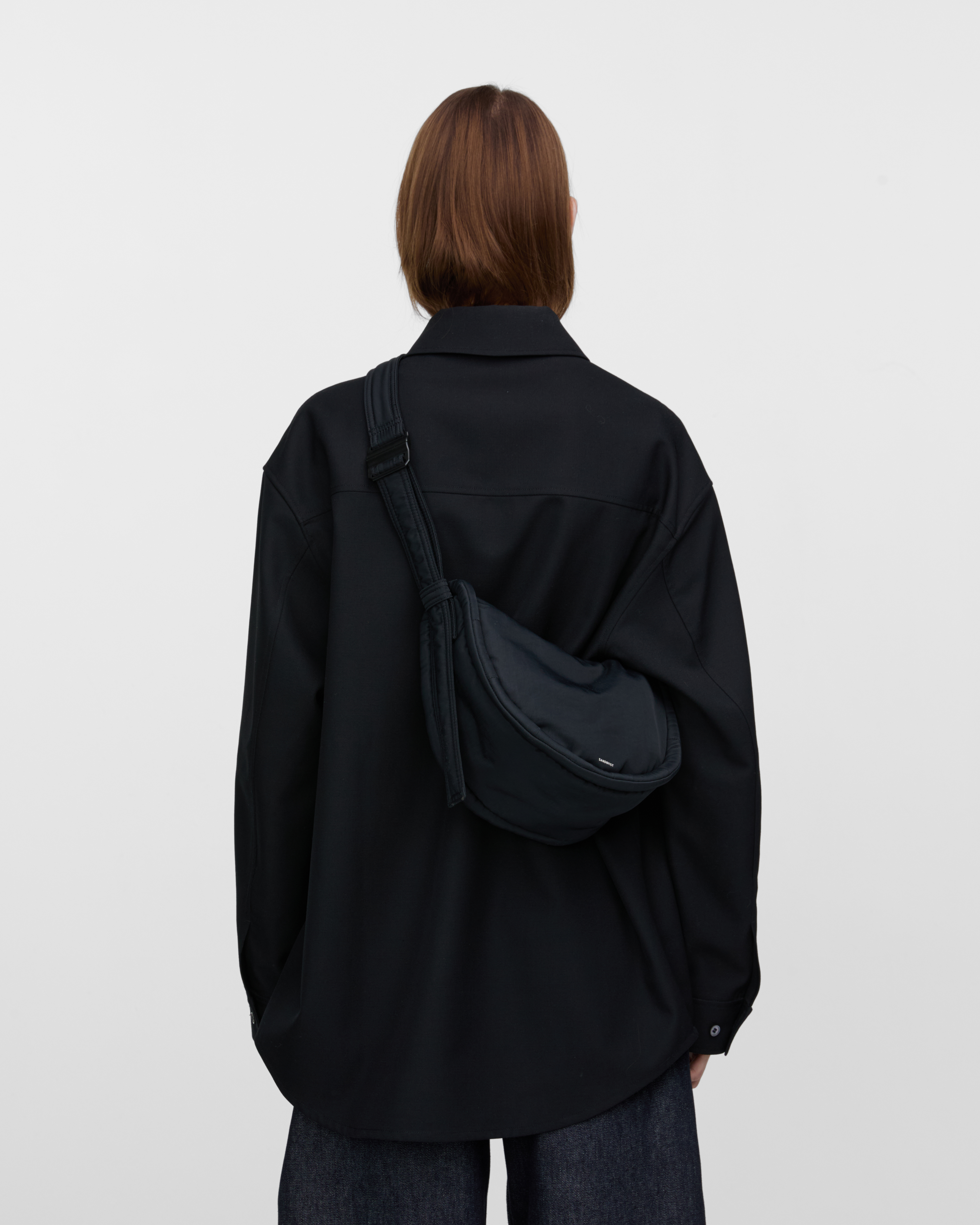 Female model wearing Sandqvist Curve Half Moon Bag Mini, black. Back view, carrying the bag on one shoulder crossbody with the bag on the back.