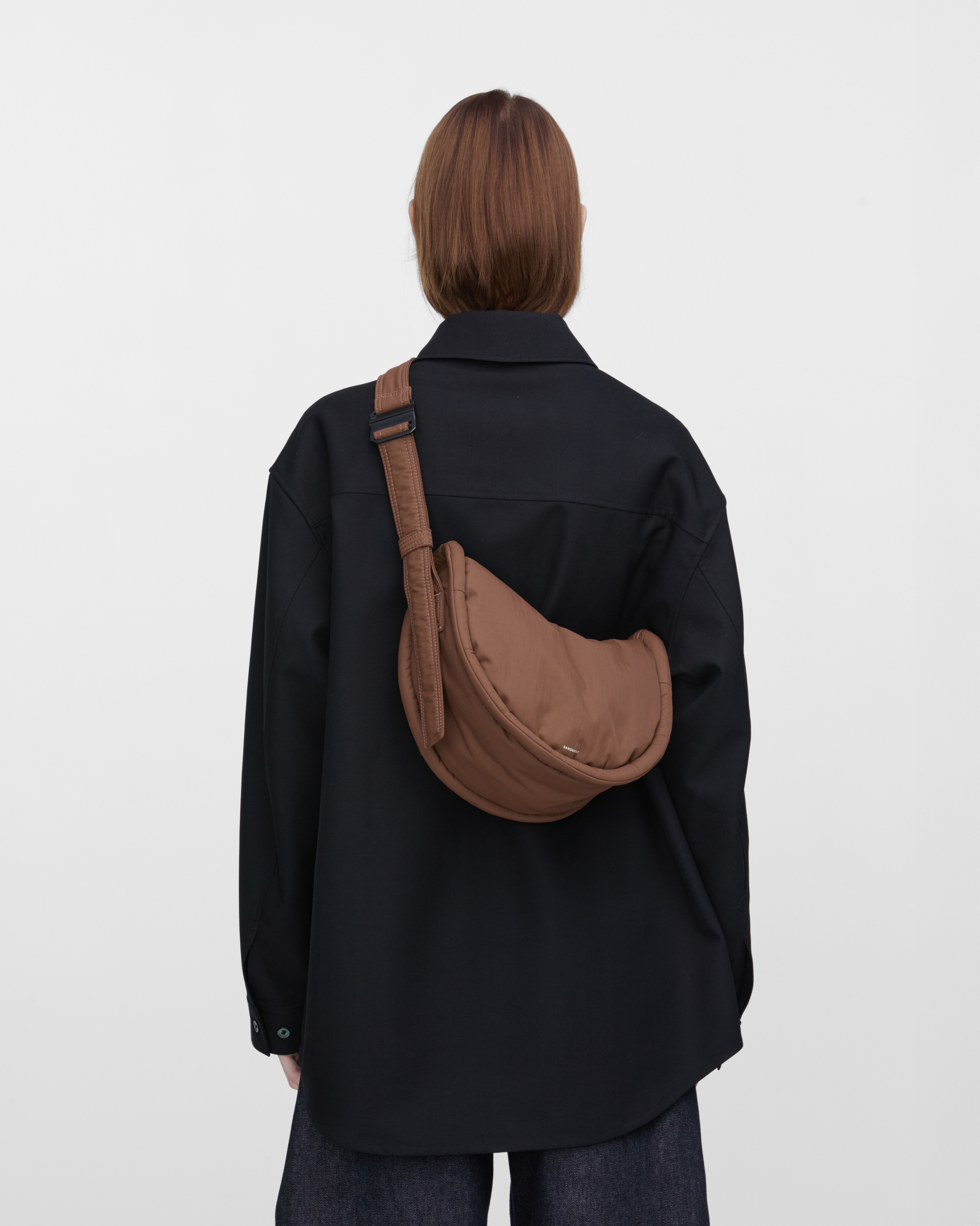 Female model wearing Sandqvist Curve Half Moon Bag Mini, brown. Back view, carrying the bag on one shoulder crossbody with the bag on the back.