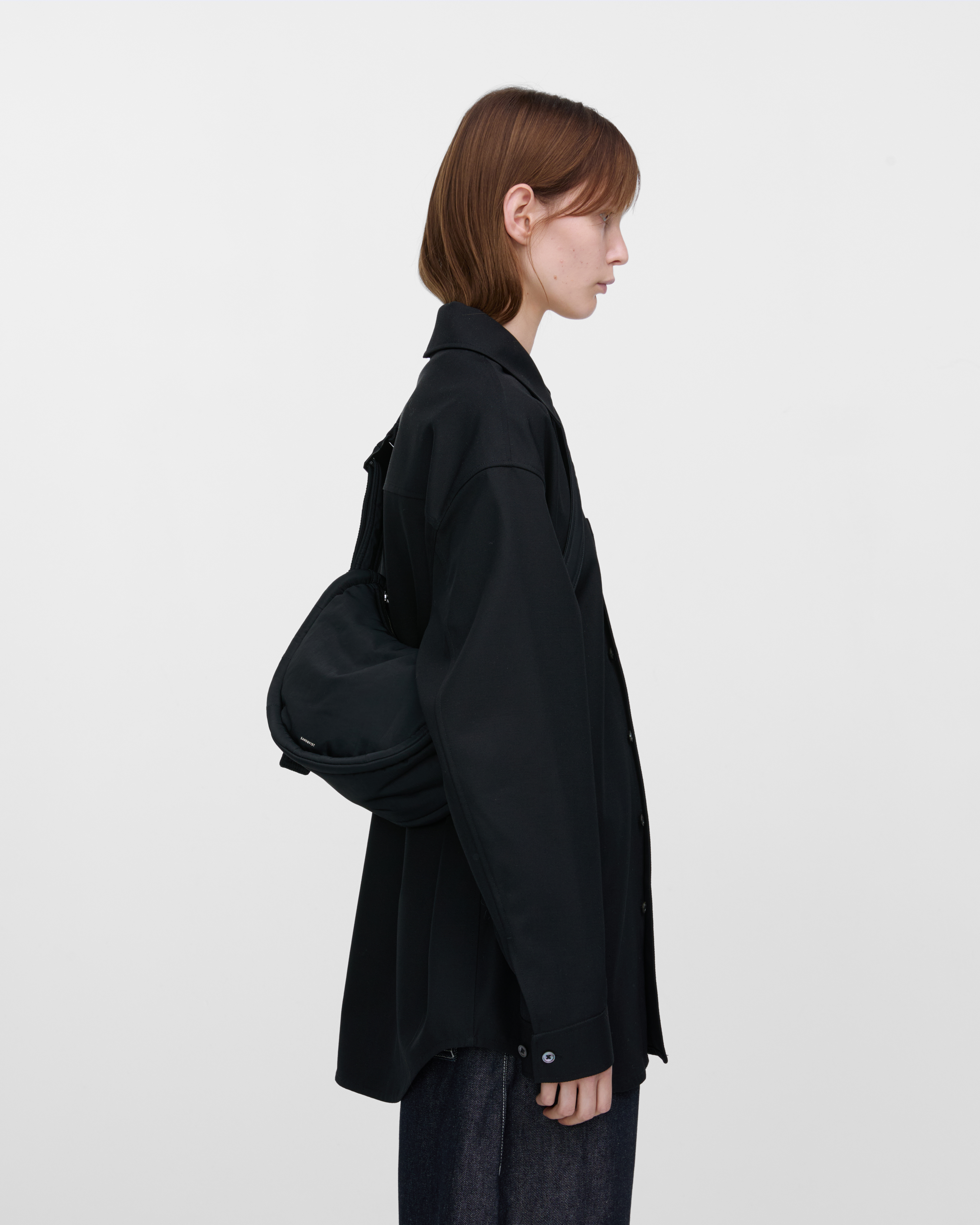 Female model wearing Sandqvist Curve Half Moon Bag Mini, black. Side view, carrying the bag on one shoulder crossbody with the bag on the back.