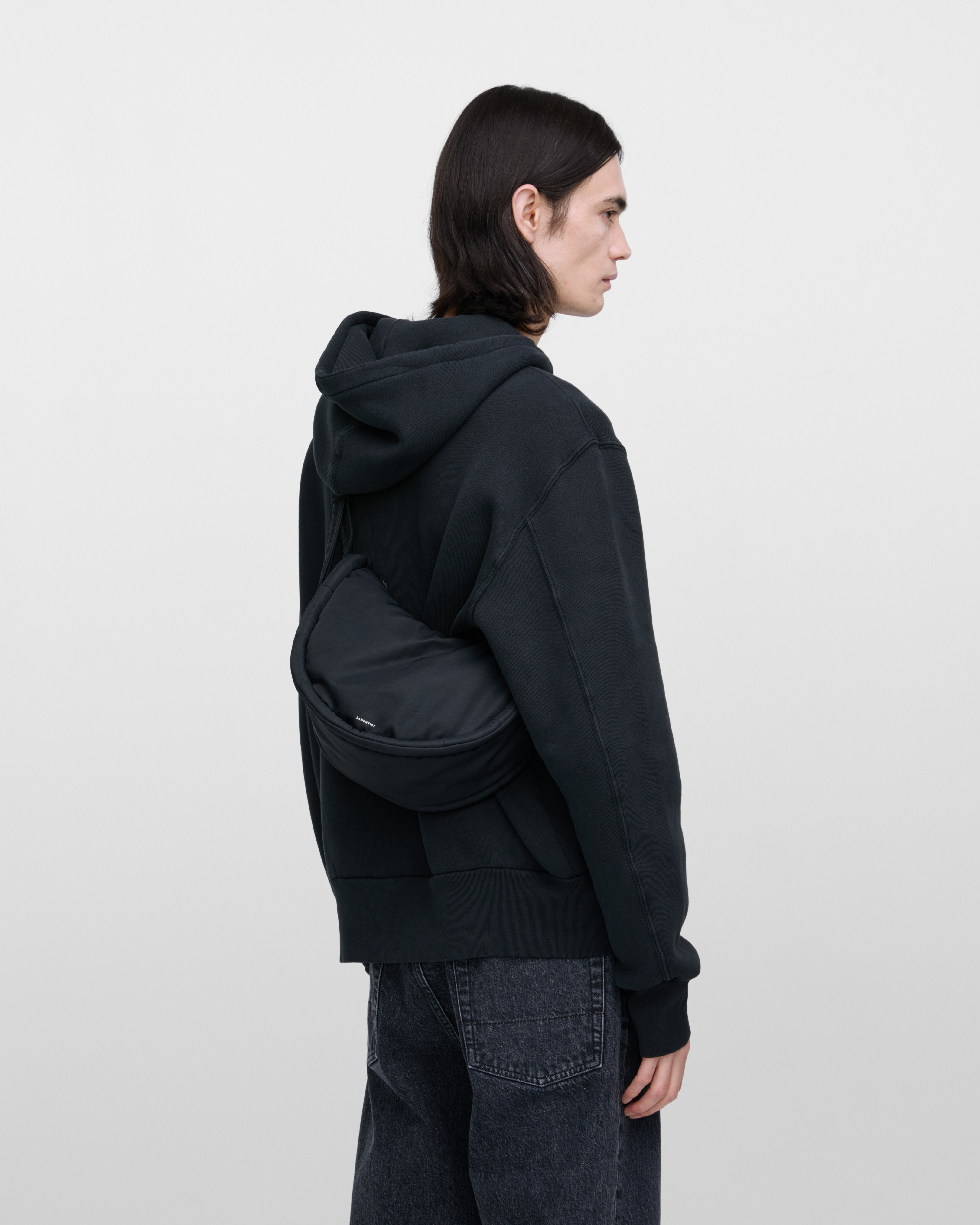 Male model wearing Sandqvist Curve Half Moon Bag Mini, black. Side view, carrying the bag on one shoulder crossbody with the bag on the back.