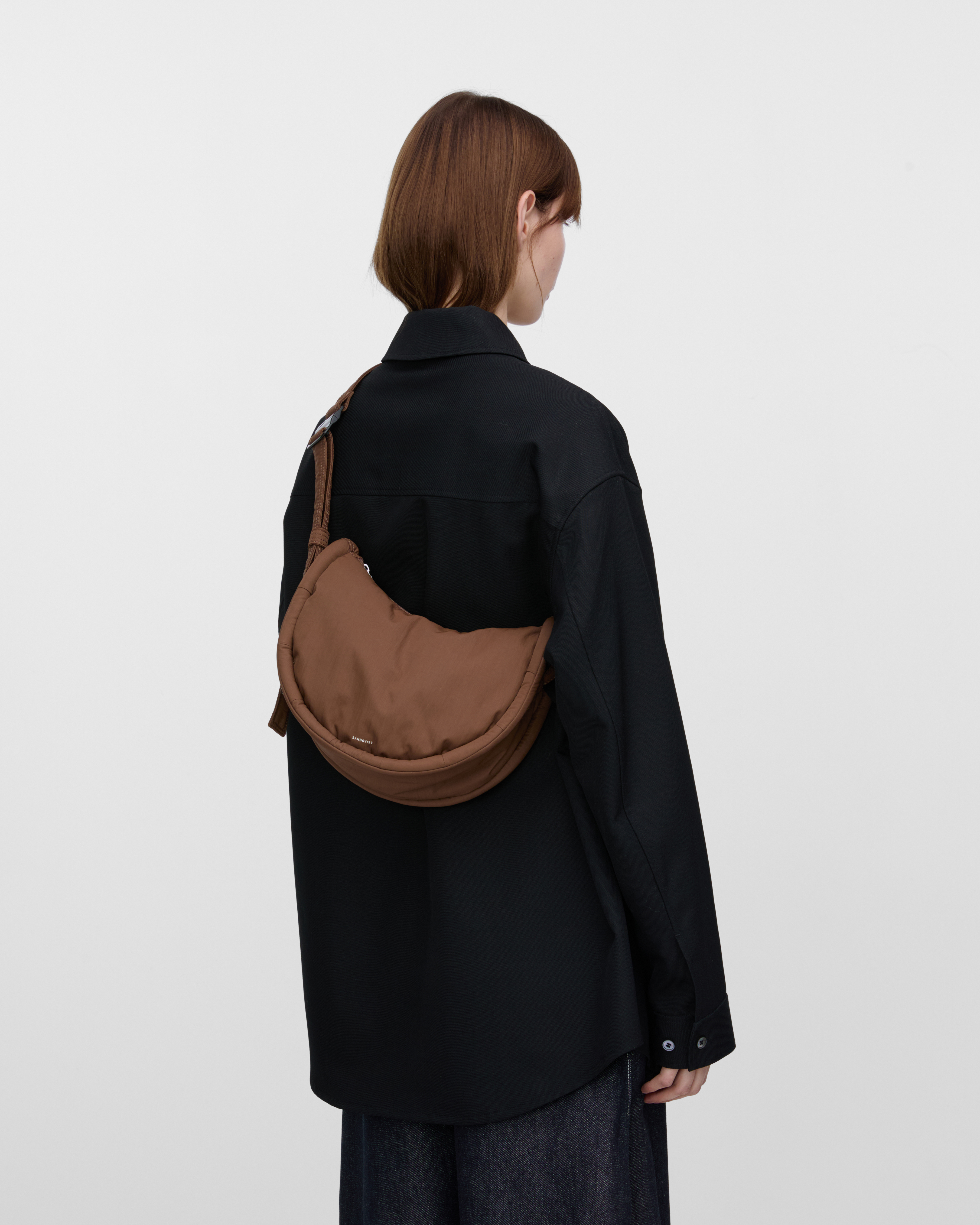 Female model wearing Sandqvist Curve Half Moon Bag Mini, brown. Back view, carrying the bag on one shoulder crossbody with the bag on the back.