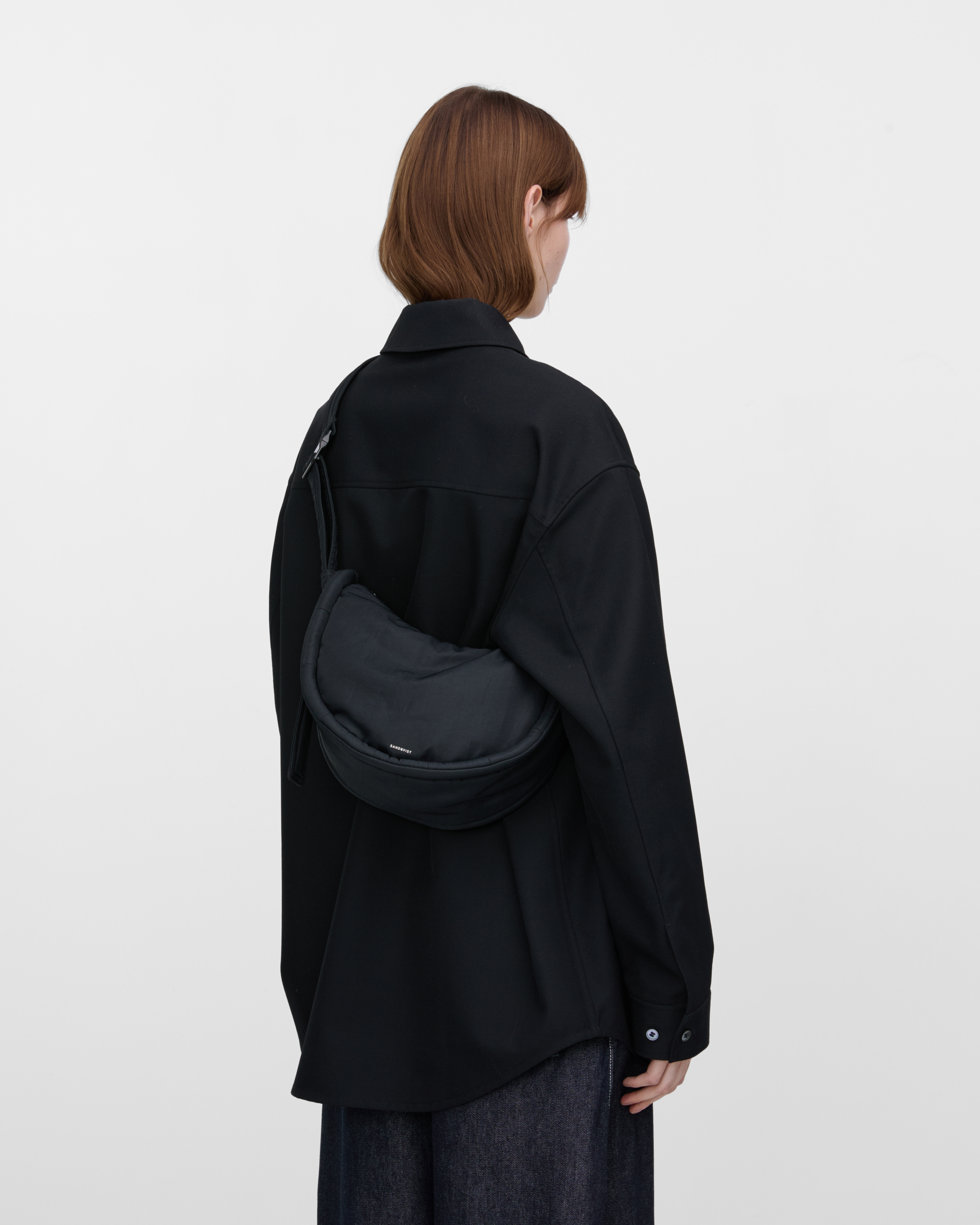 Female model wearing Sandqvist Curve Half Moon Bag Mini, black. Back view, carrying the bag on one shoulder crossbody with the bag on the back.