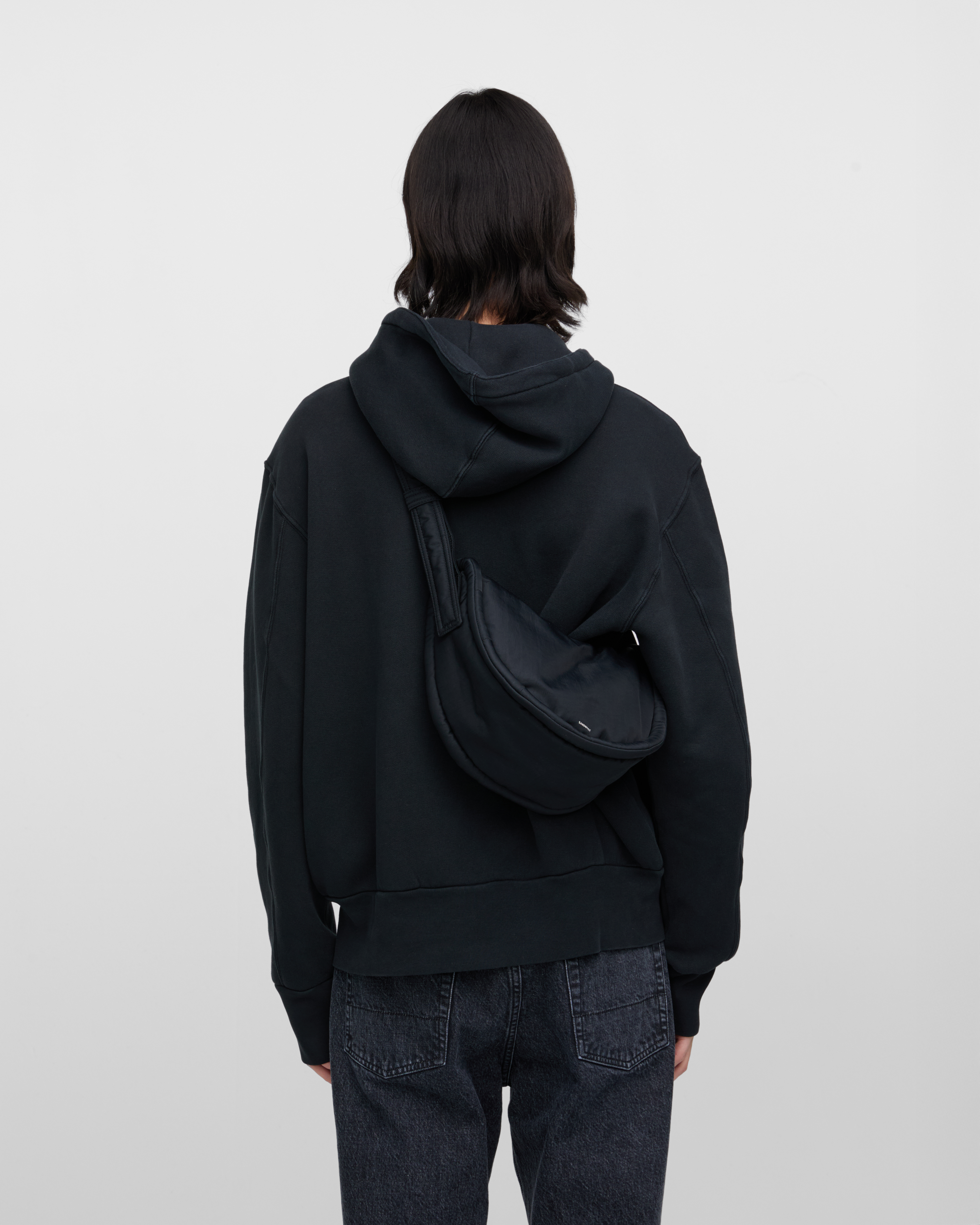 Male model wearing Sandqvist Curve Half Moon Bag Mini, black. Back view, carrying the bag on one shoulder crossbody with the bag on the back.