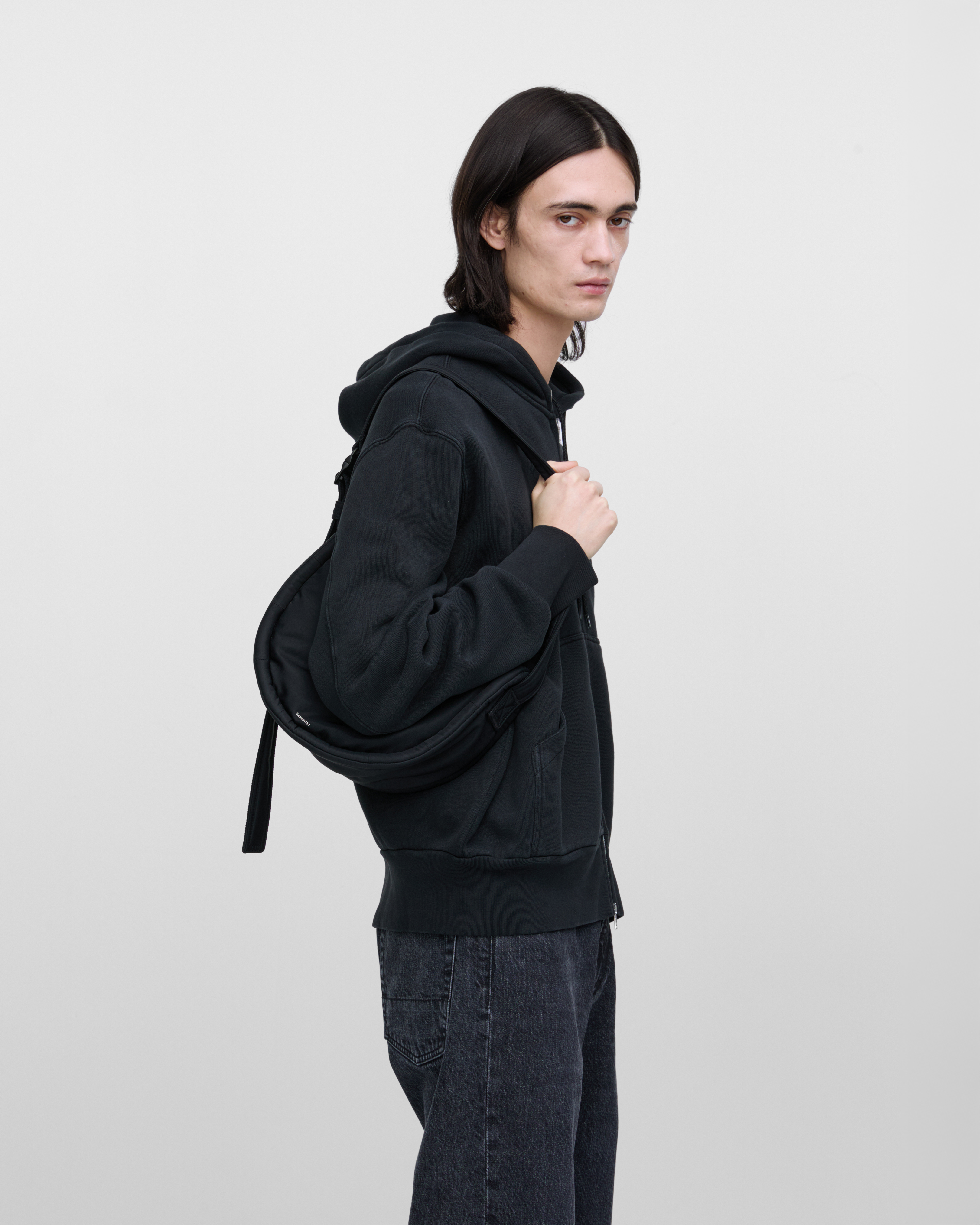 Male model wearing Sandqvist Curve Half Moon Bag Mini, black. Side view, carrying the bag on one shoulder.