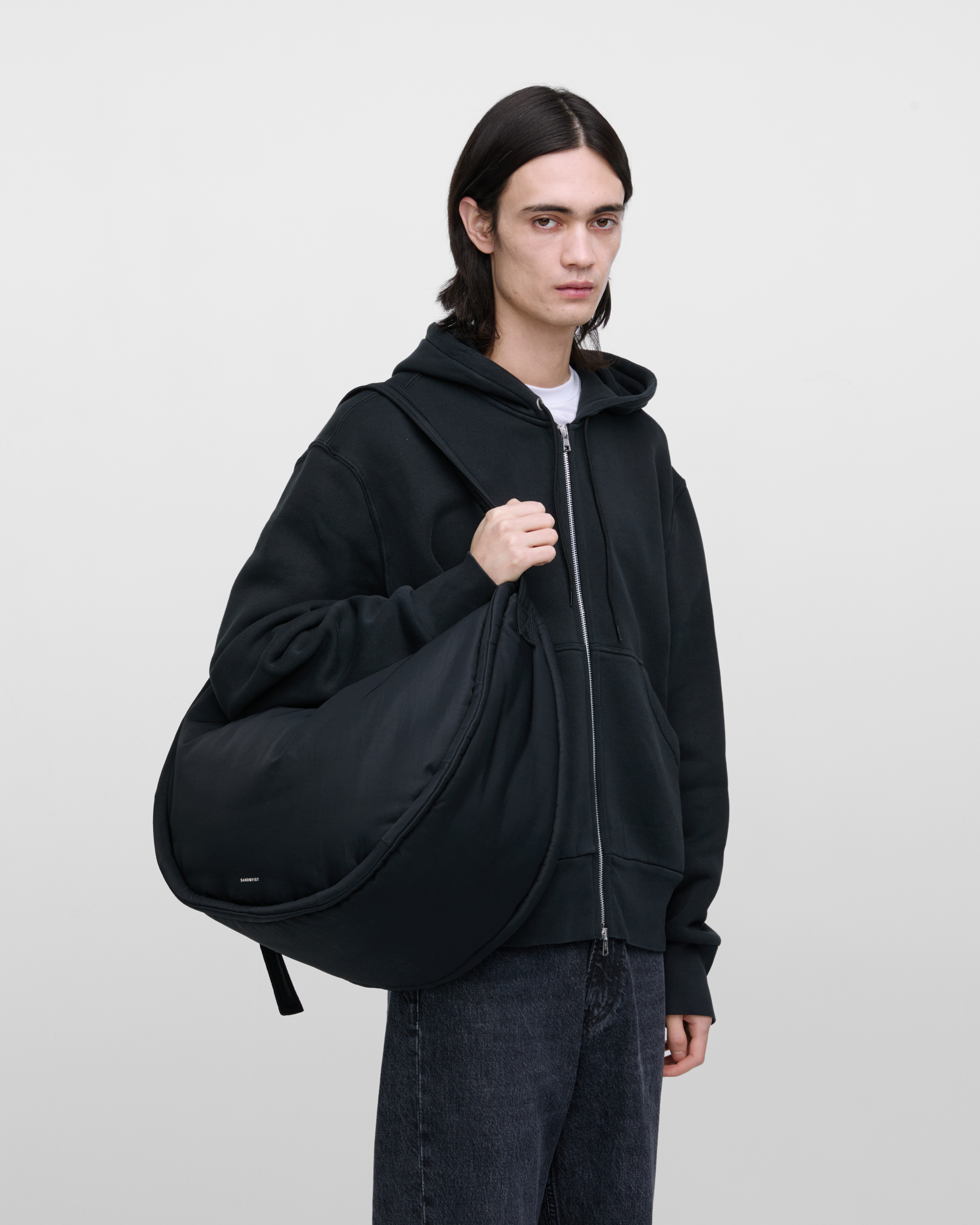 Male model carrying Sandqvist Curve Half Moon Bag, black. Front view, carrying the bag over one shoulder.