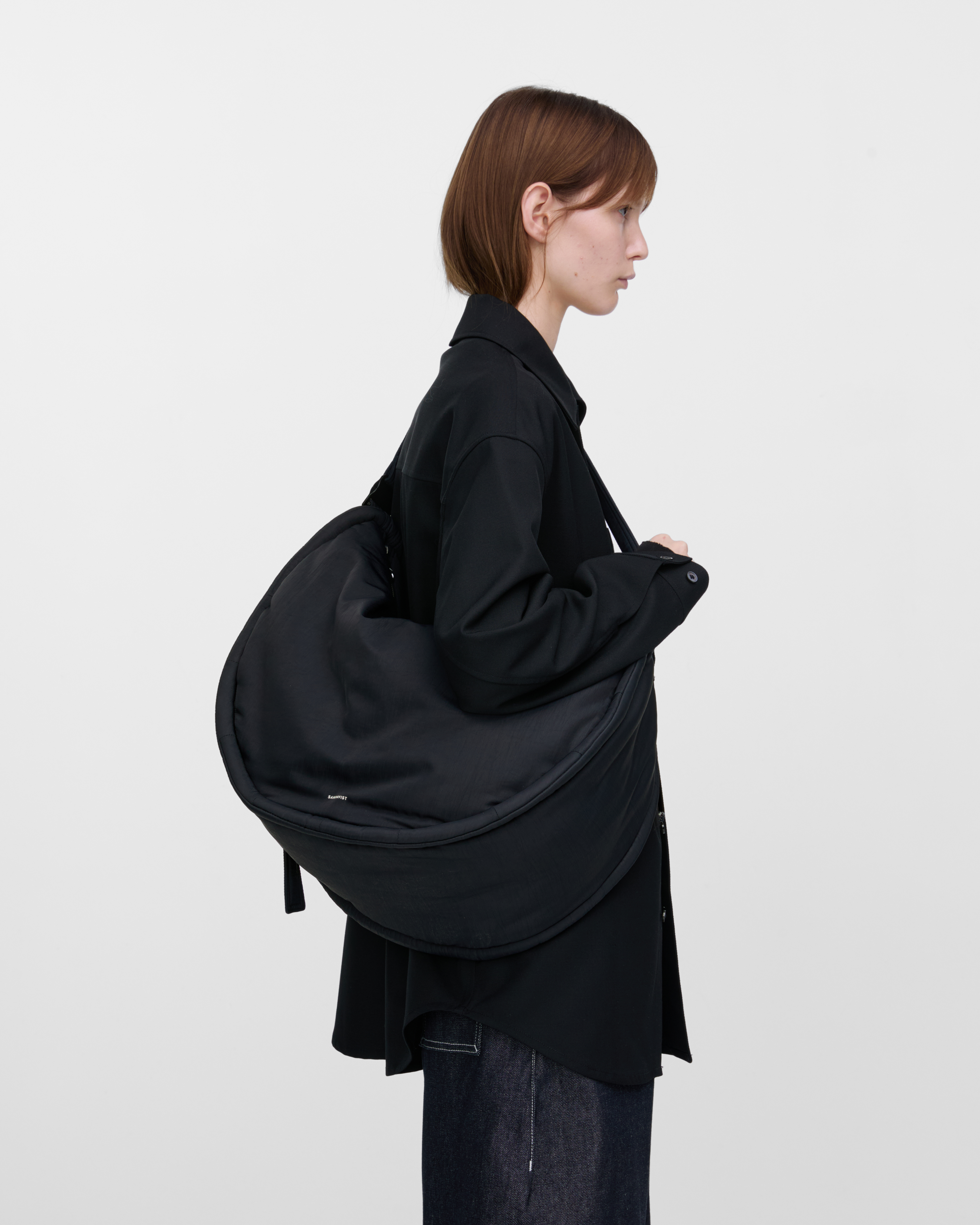 Female model wearing Sandqvist Curve Half Moon Bag, black. Side view, Wearing the bag on one shoulder crossbody.