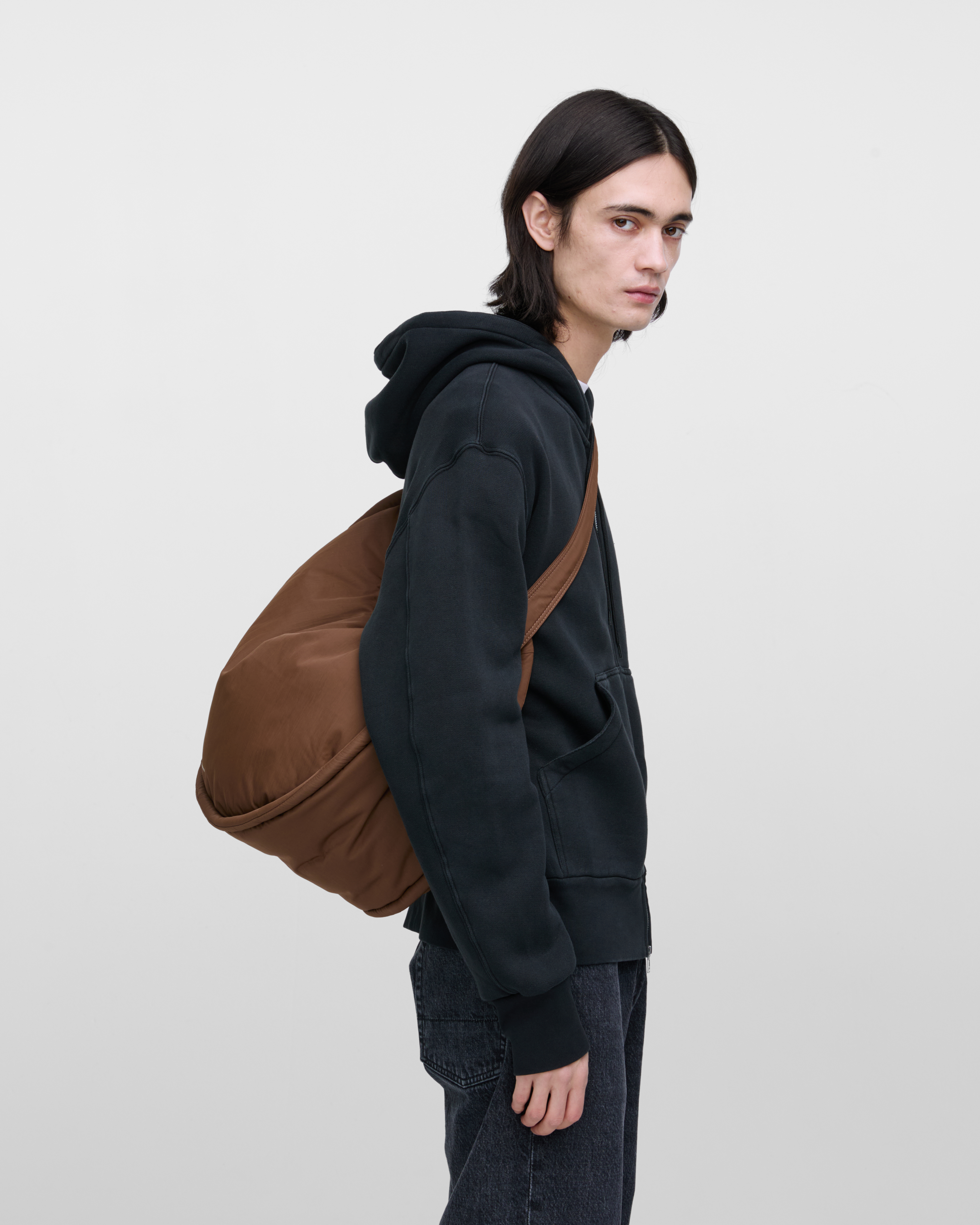 Männliches Model mit Sandqvist Curve Half Moon Bag, braun. Seitenansicht, die Tasche über einer Schulter quer über den Körper getragen.