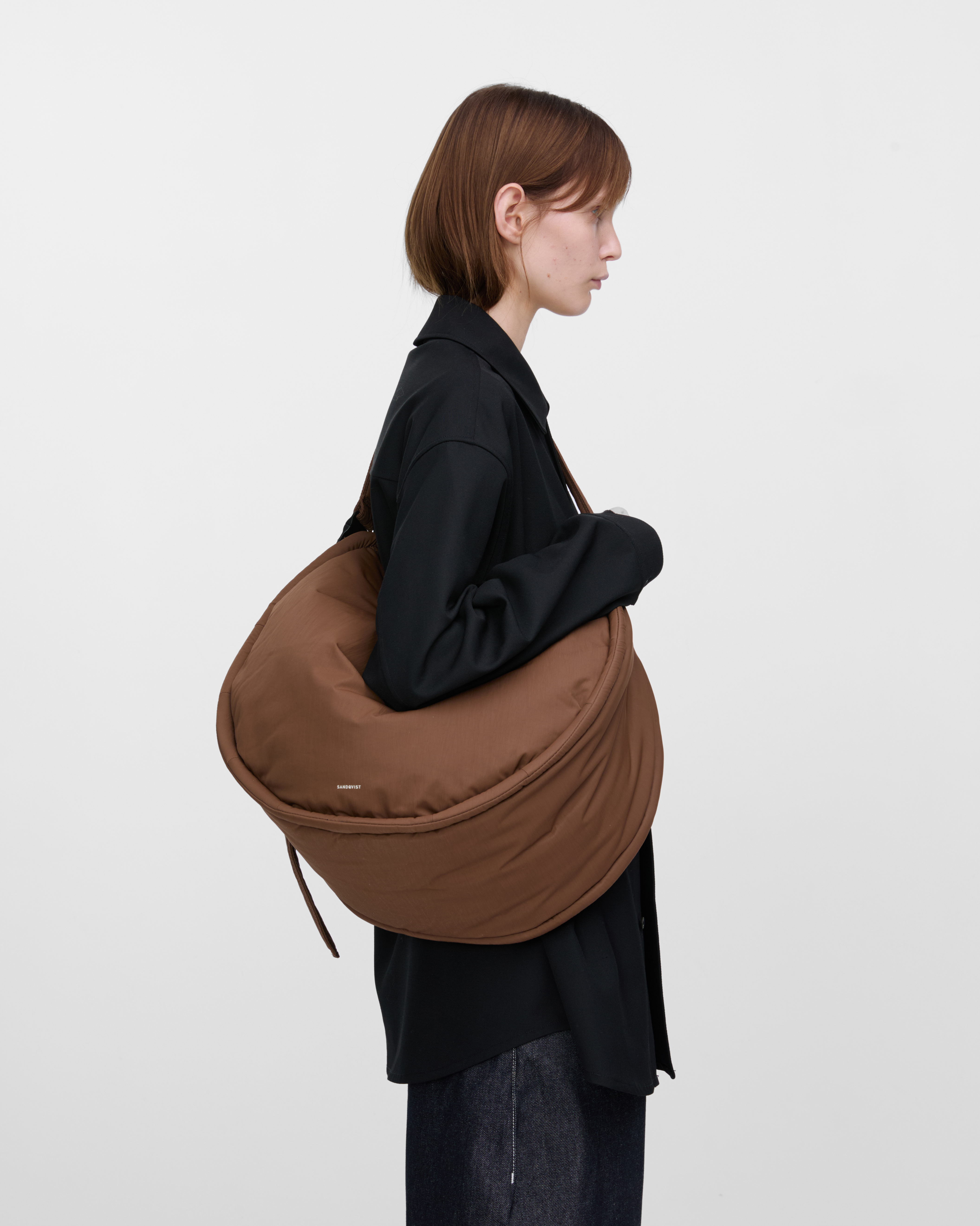 Weibliches Model mit Sandqvist Curve Half Moon Bag, braun. Seitenansicht, die Tasche über einer Schulter quer über den Körper getragen.