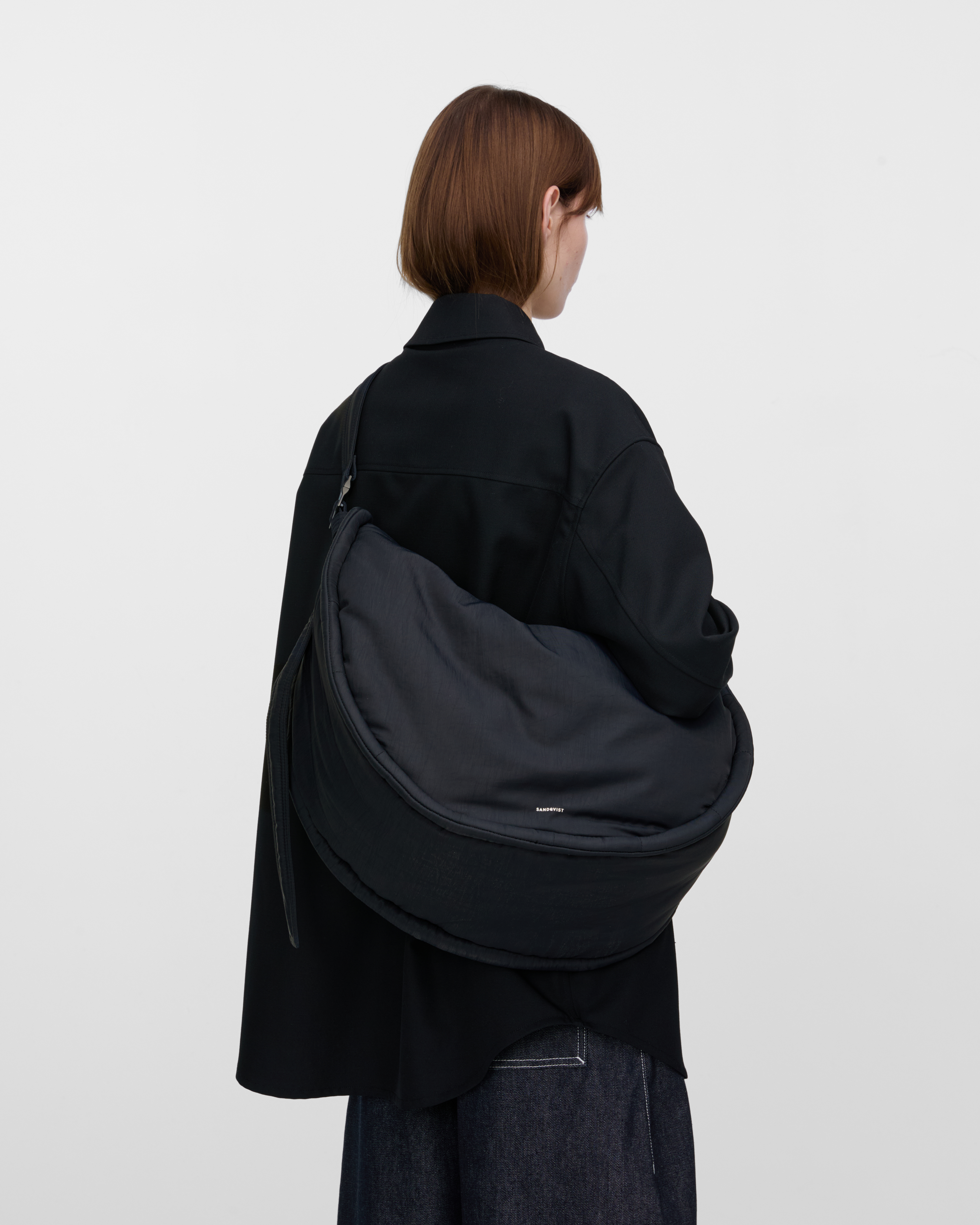 Female model wearing Sandqvist Curve Half Moon Bag, black. Back view, carrying the bag on one shoulder crossbody.