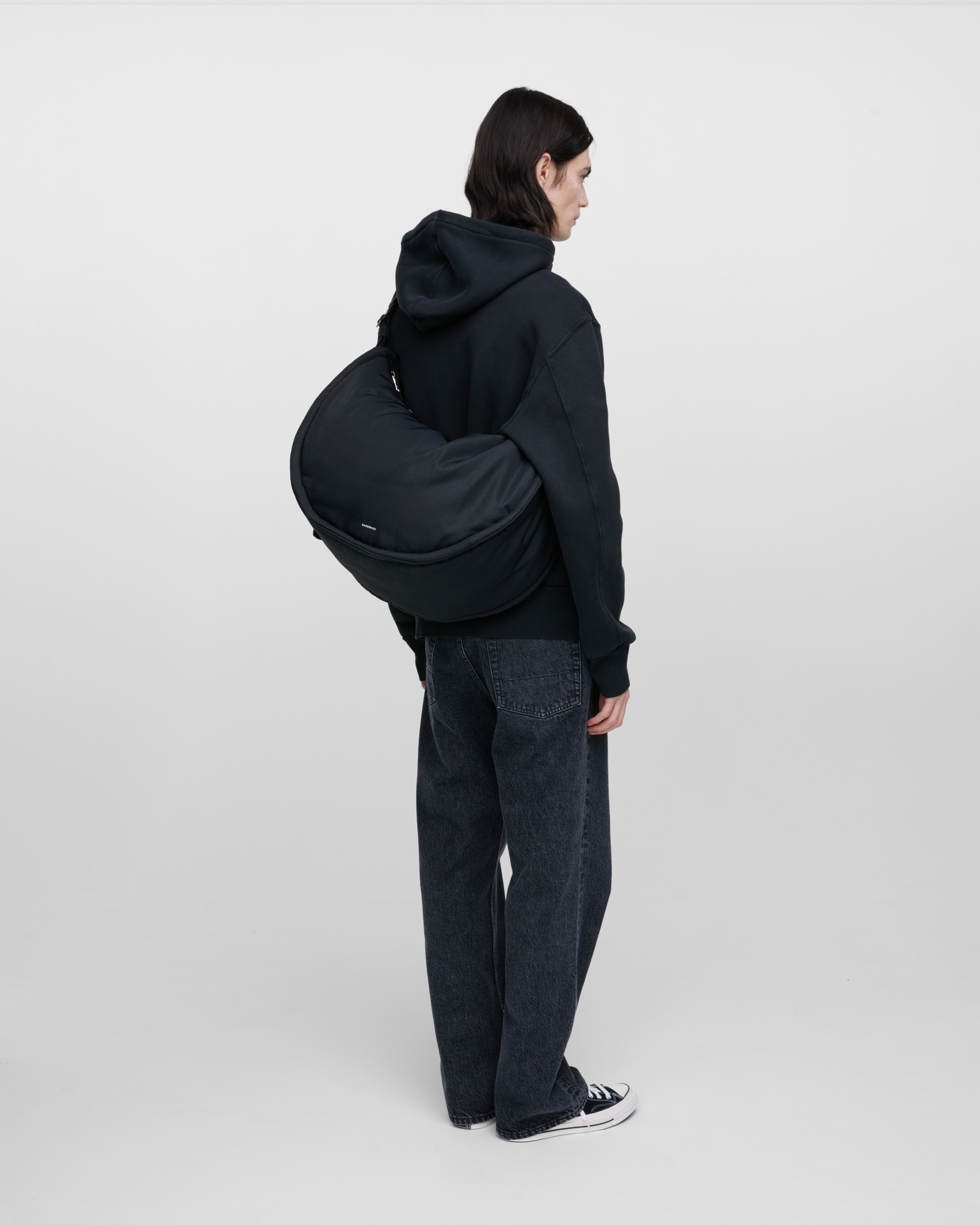 Male model carrying Sandqvist Curve Half Moon Bag, black. Back view, carrying the bag over one shoulder crossbody, with the bag in the back.