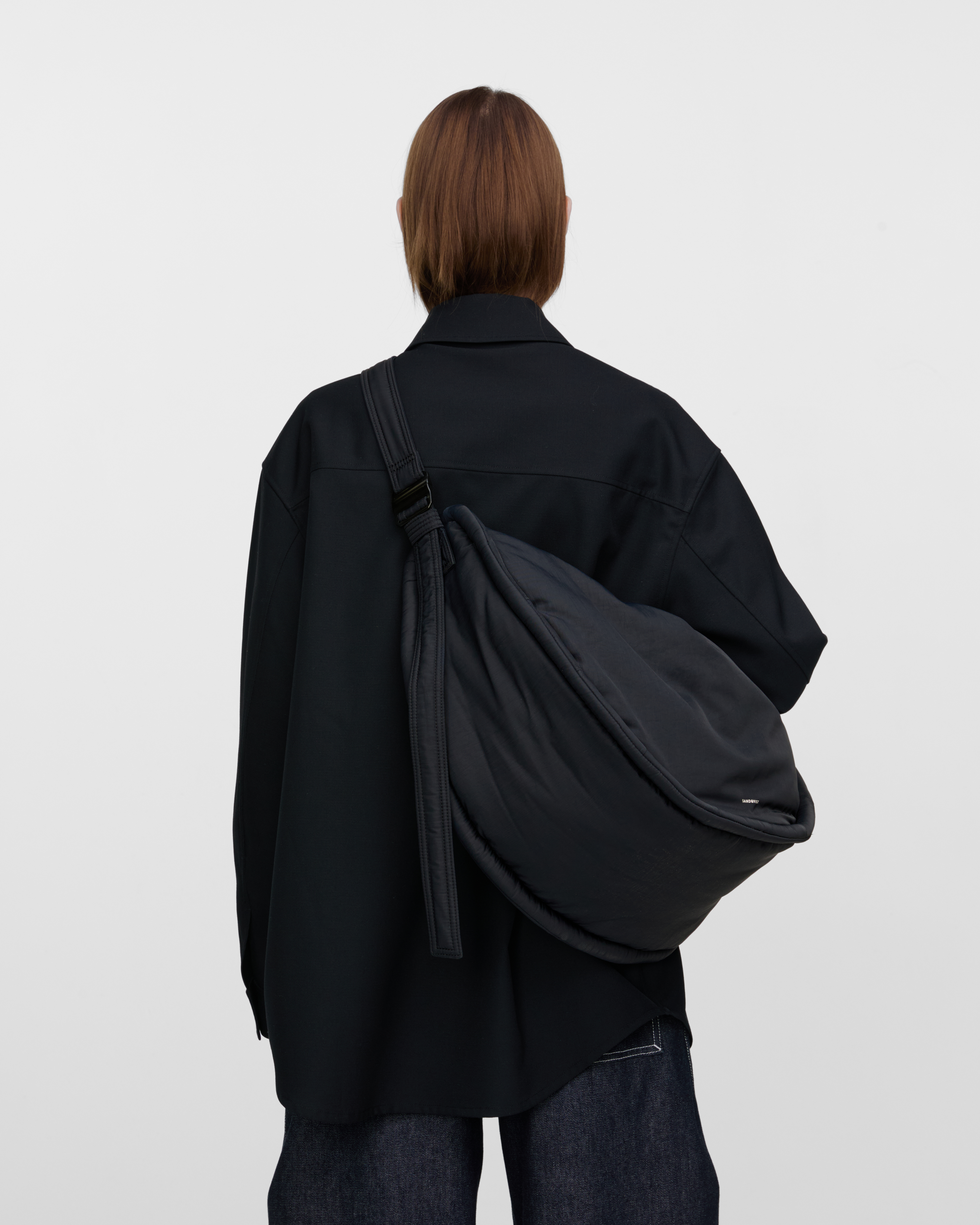 Female model wearing Sandqvist Curve Half Moon Bag, black. Back view, carrying the bag on one shoulder crossbody with the bag on the back.