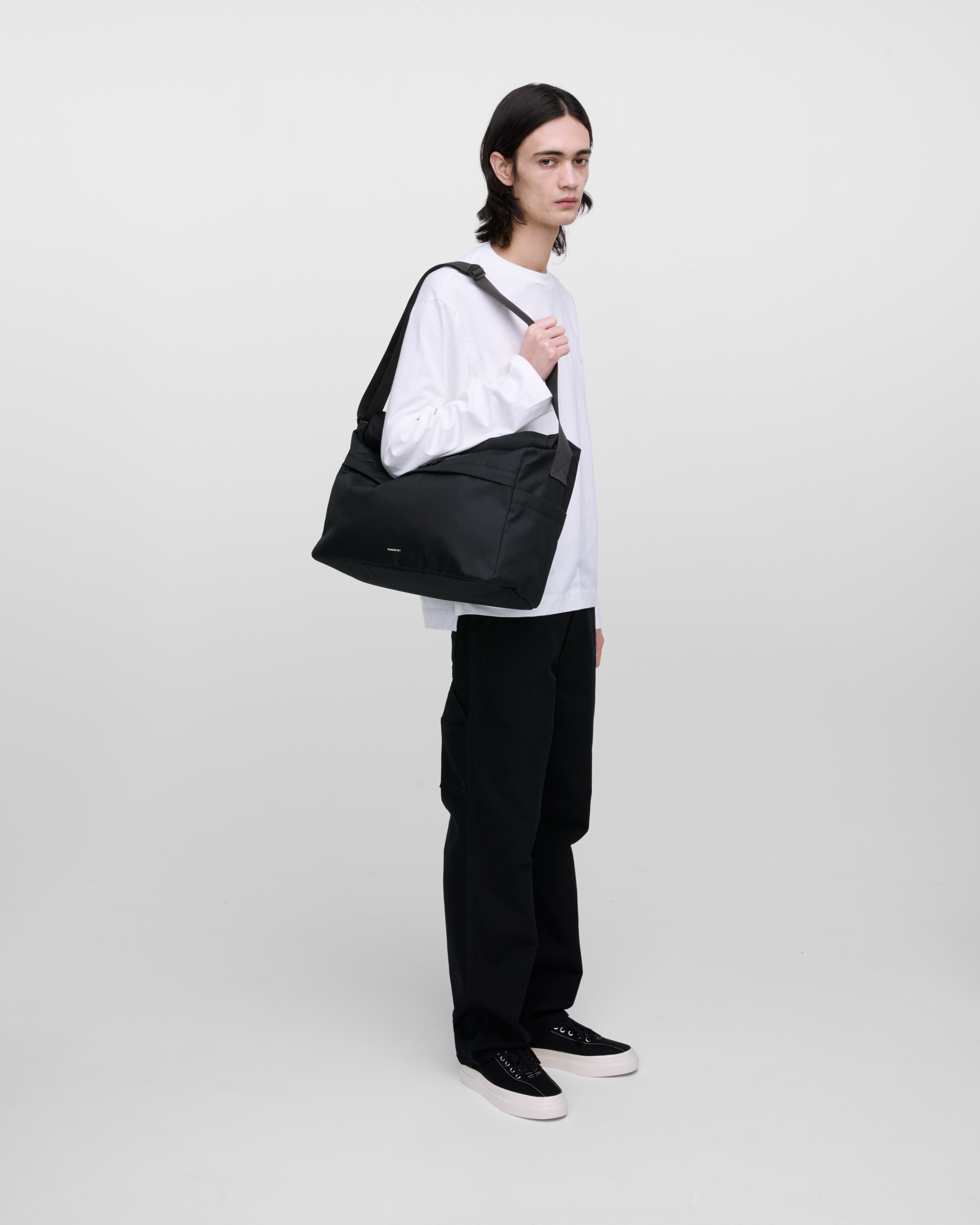 Male model wearing Sandqvist Icon Messenger Bag, black. Side view, wearing the bag on one shoulder.