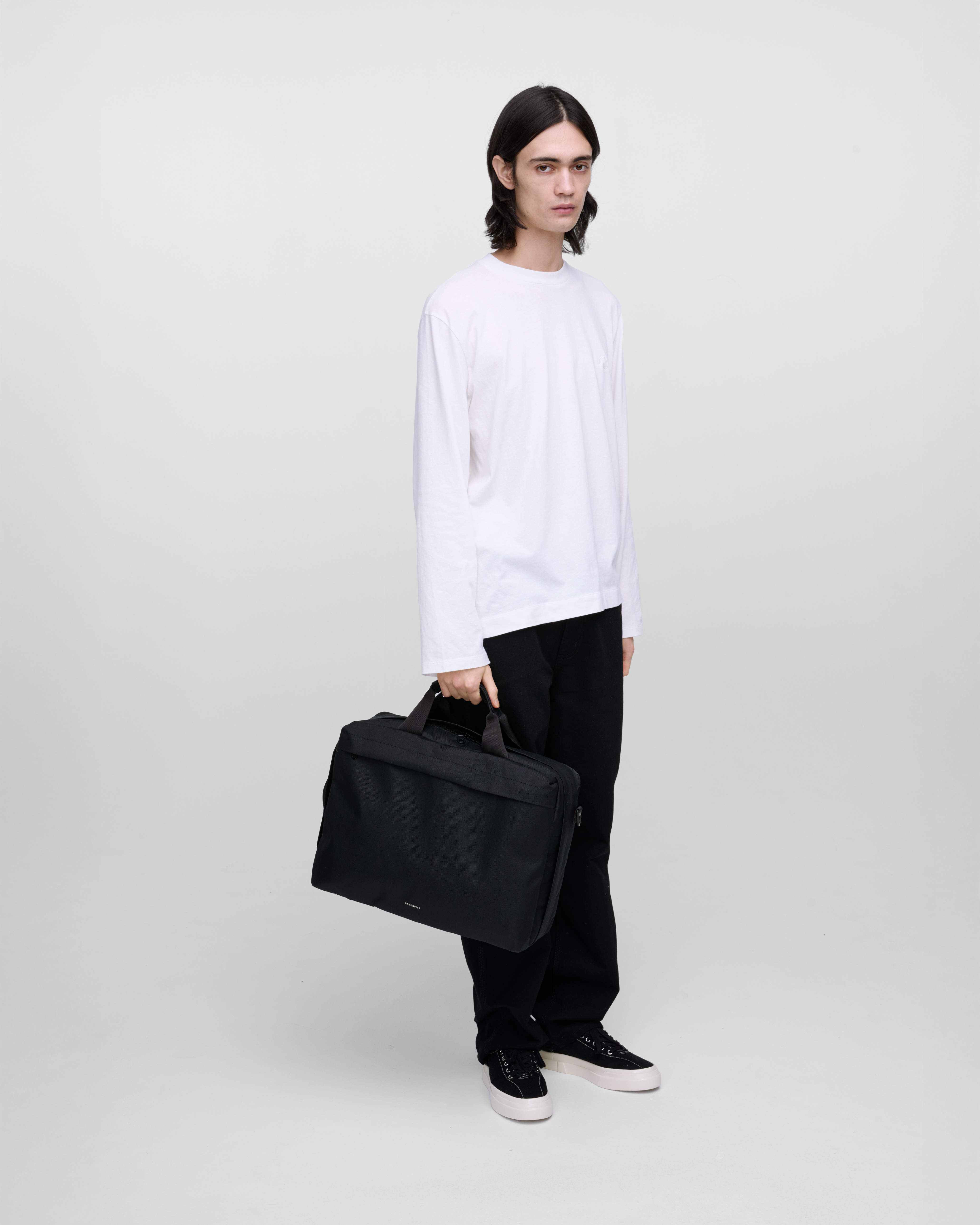 Male model holding Sandqvist GO 3-Way Bag M, black. Front view, holding the bag in one hand, in the top handle.