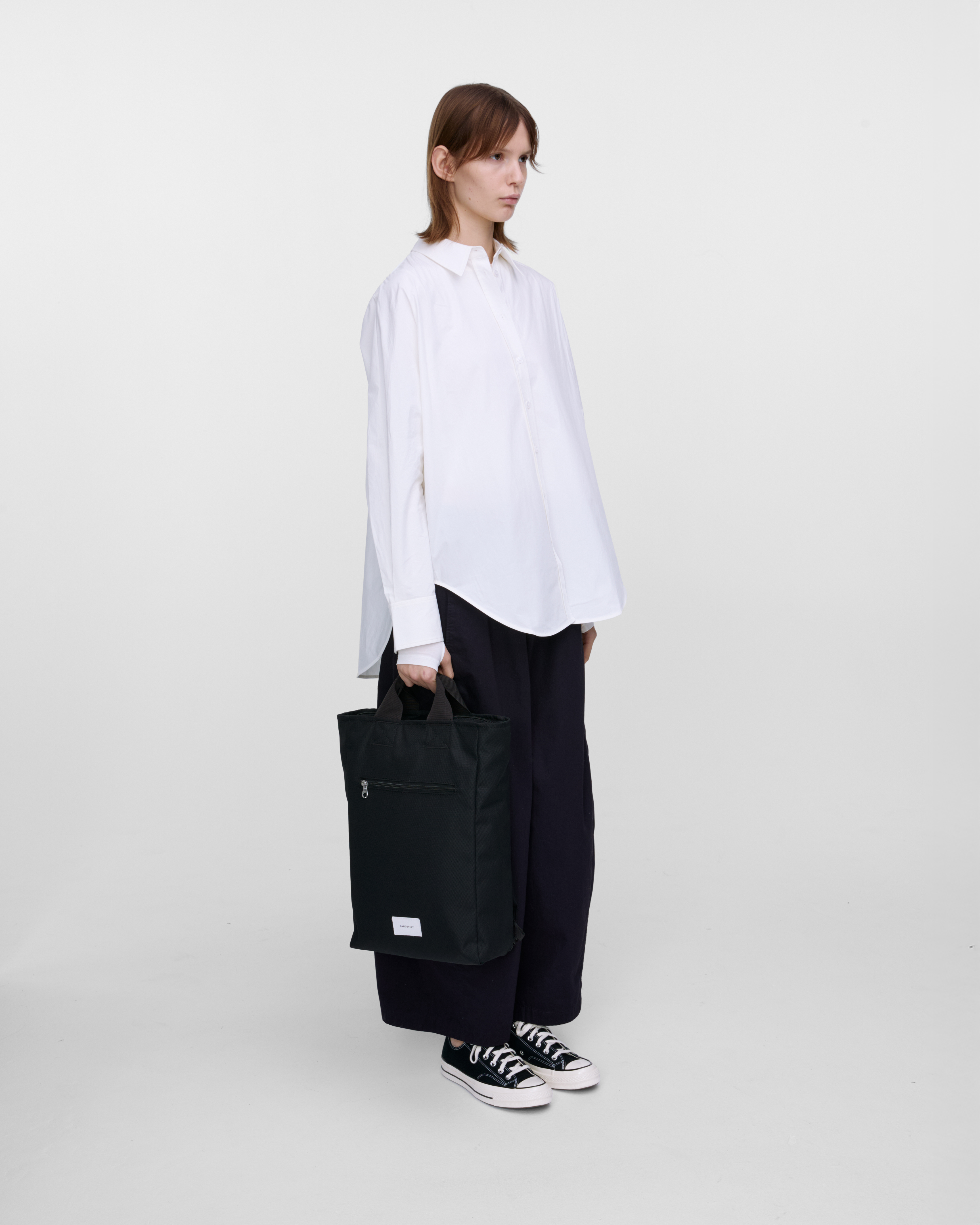 Female model holding Sandqvist Ground 2-way Bag, black. Side view, holding the bag in one hand,