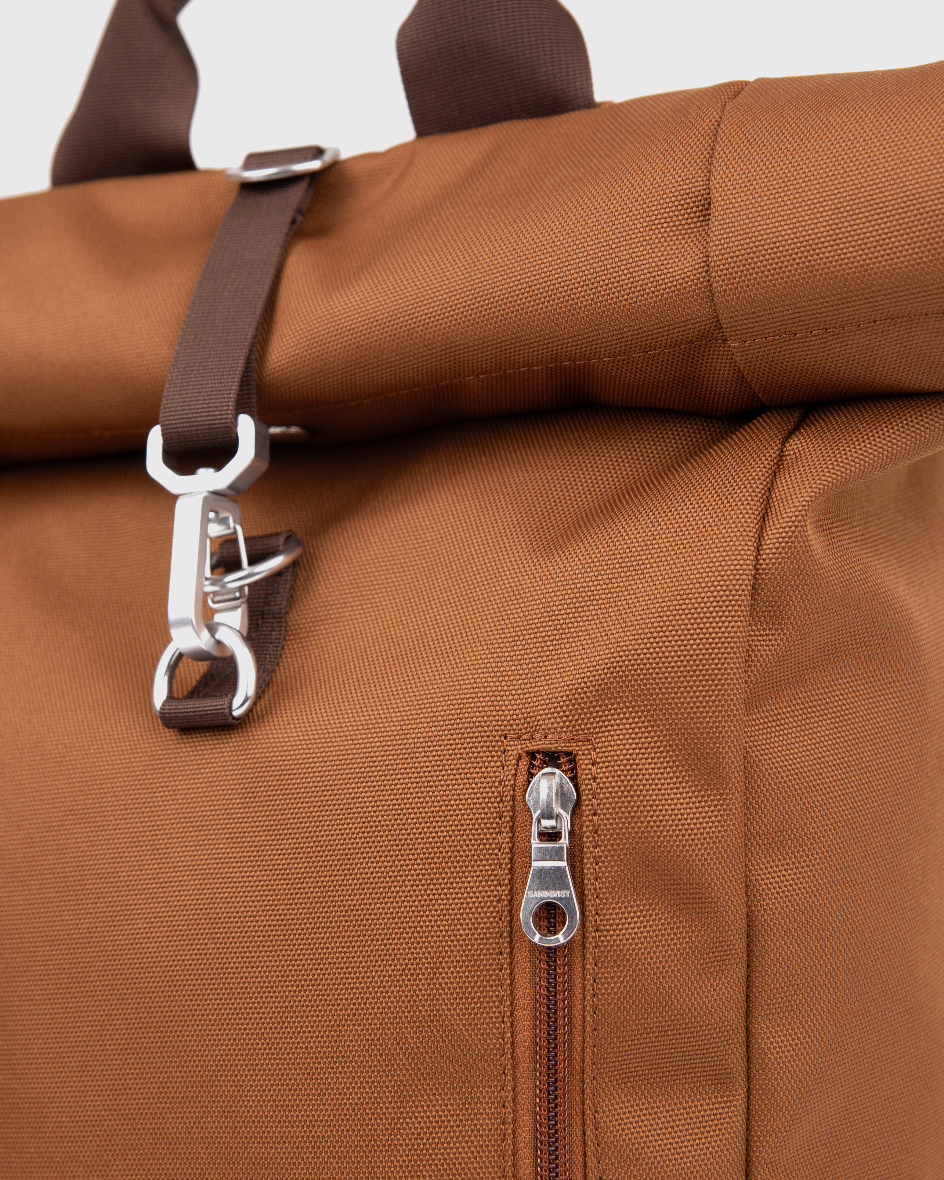 Sandqvist Ground Rolltop Backpack L, brown. Close-up of front buckle and zipper of the front pocket.