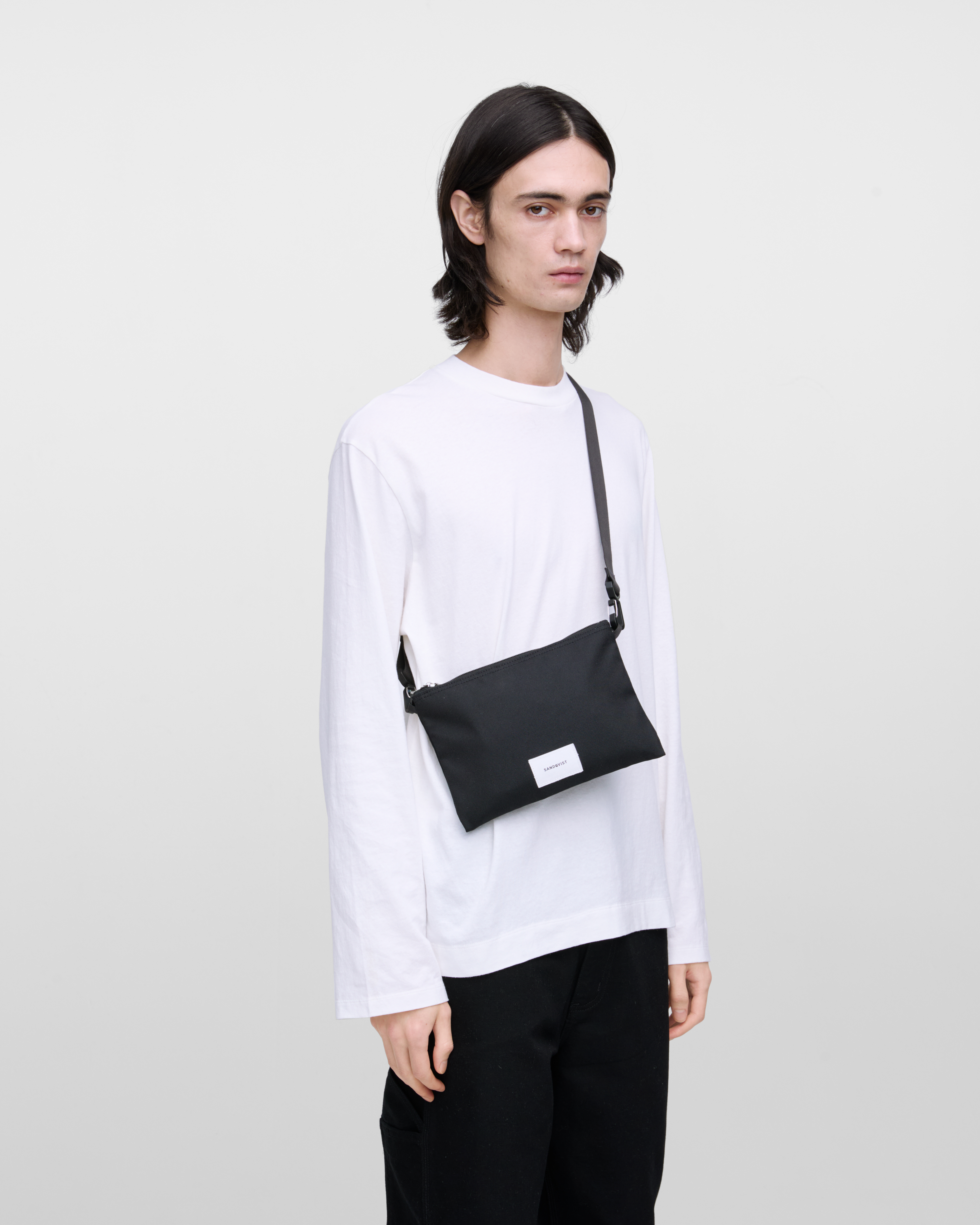 Male model wearing Sandqvist Ground Sacoche Slim, black. Front view, wearing the bag on one shoulder crossbody.