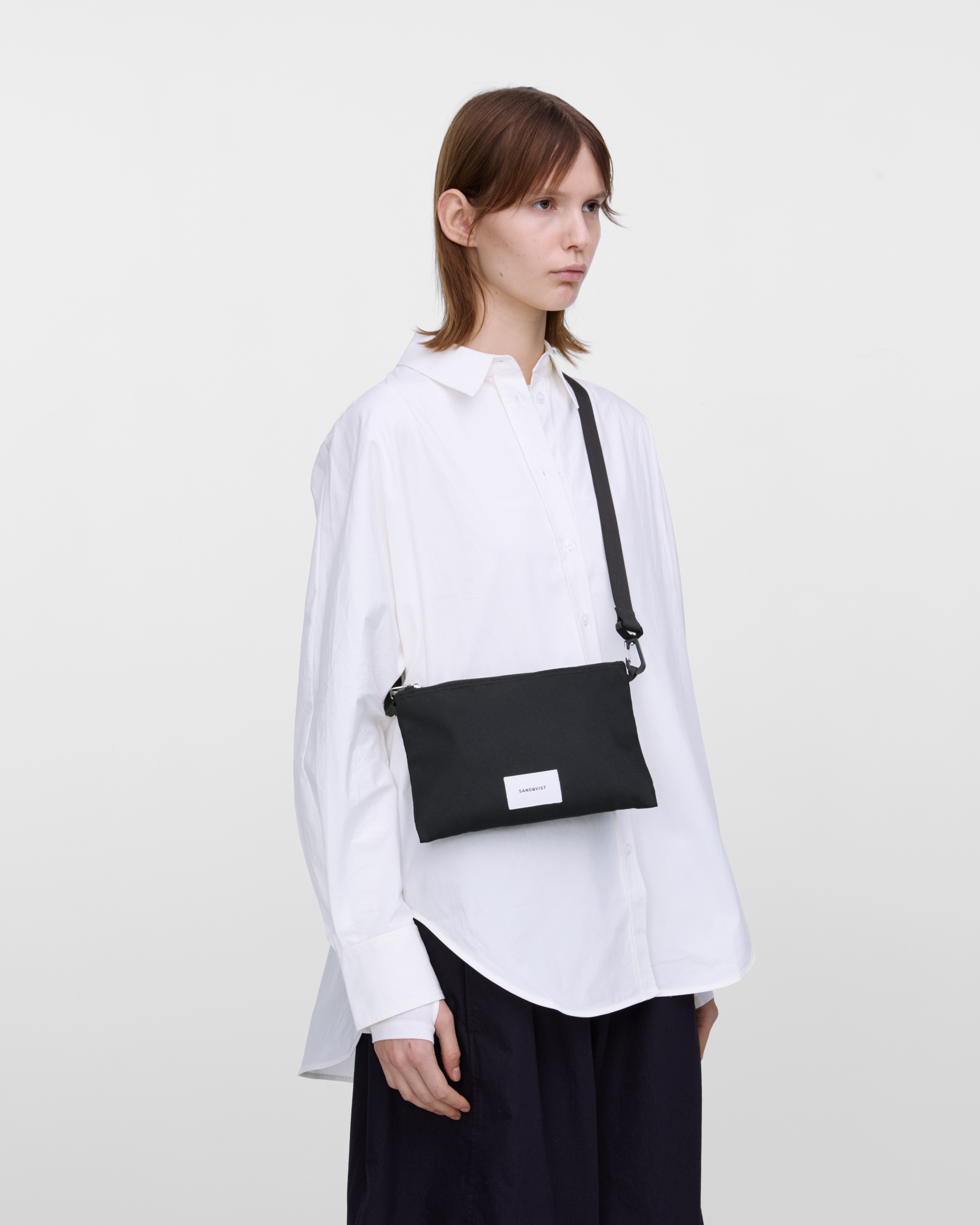 Female model wearing Sandqvist Ground Sacoche Slim, black. Front view, wearing the bag on one shoulder crossbody.