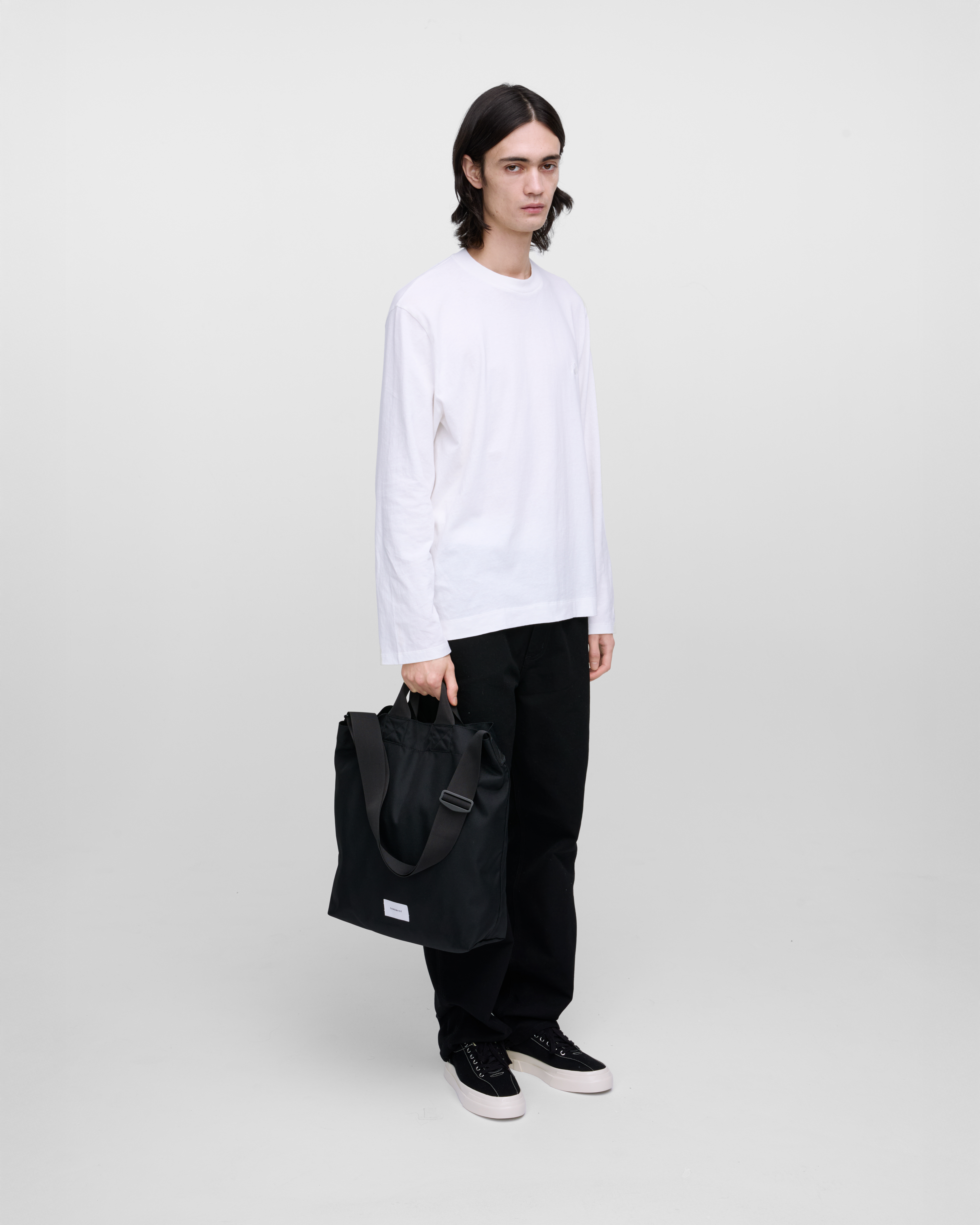 Male model holding Sandqvist Ground Shopper Bag, black. Front view, holding the bag in one hand.