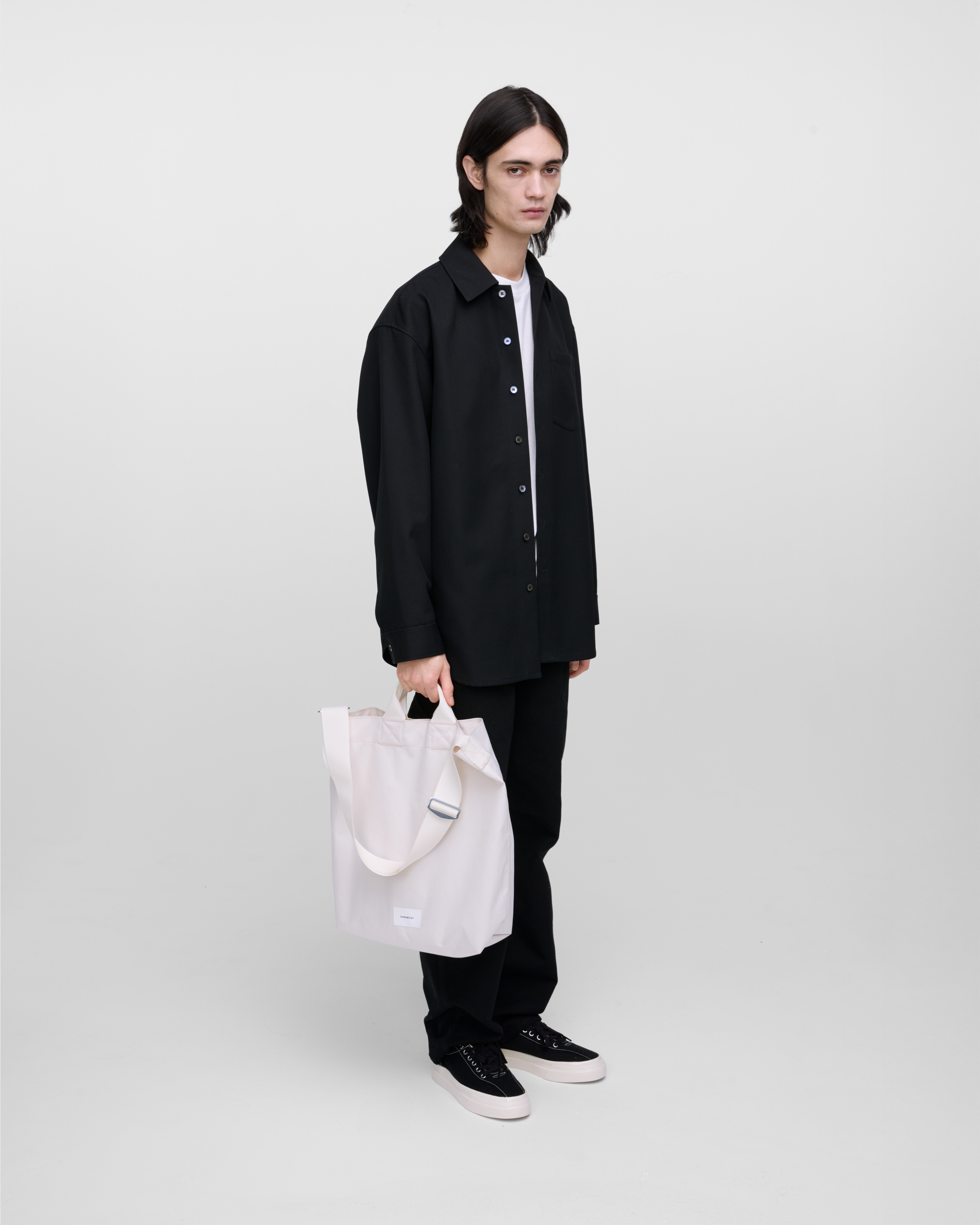 Male model holding Sandqvist Ground Shopper Bag, white. Side view, holding the bag in one hand.