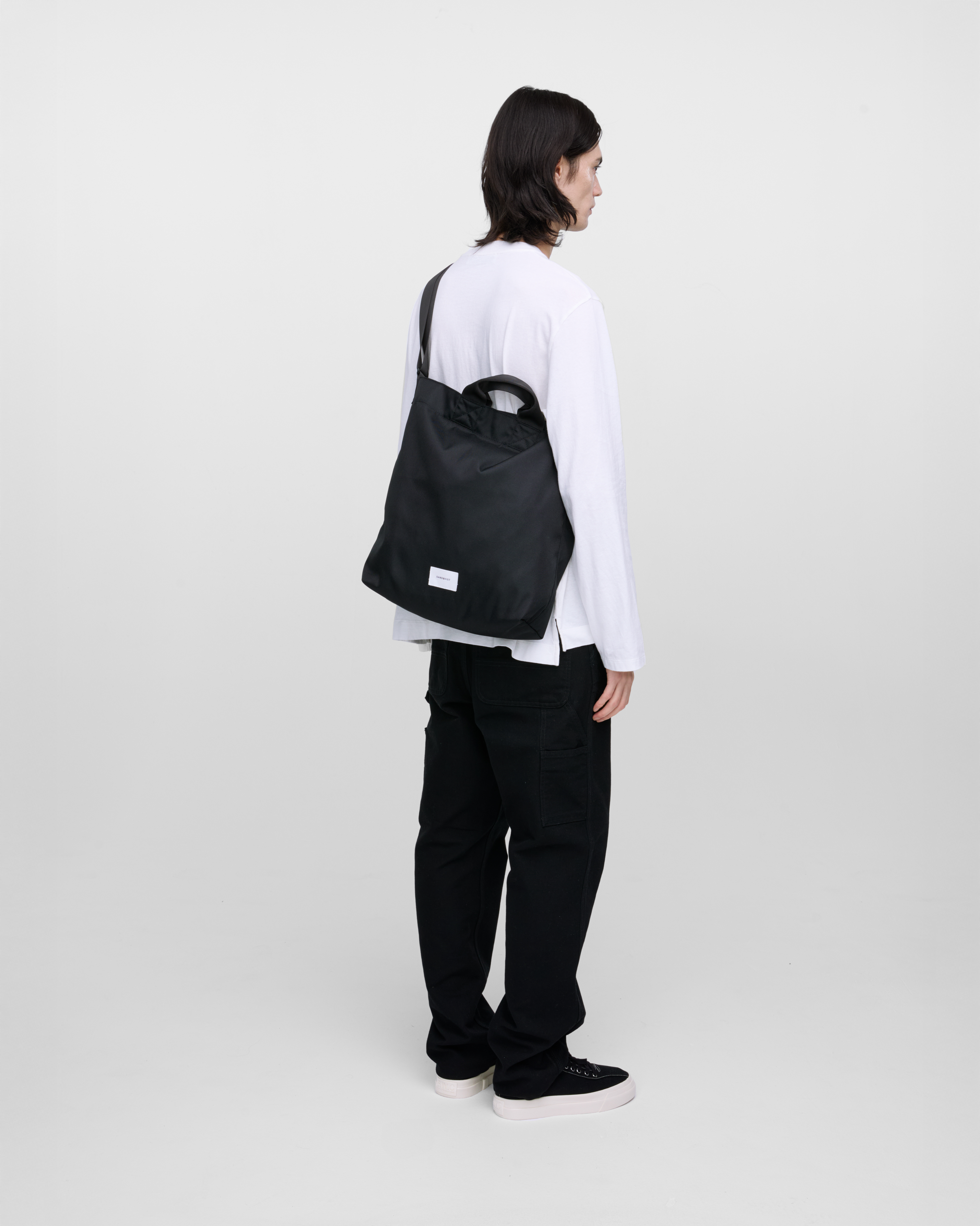 Male model wearing Sandqvist Ground Shopper Bag, black. Back view, wearing the bag on one shoulder crossbody.