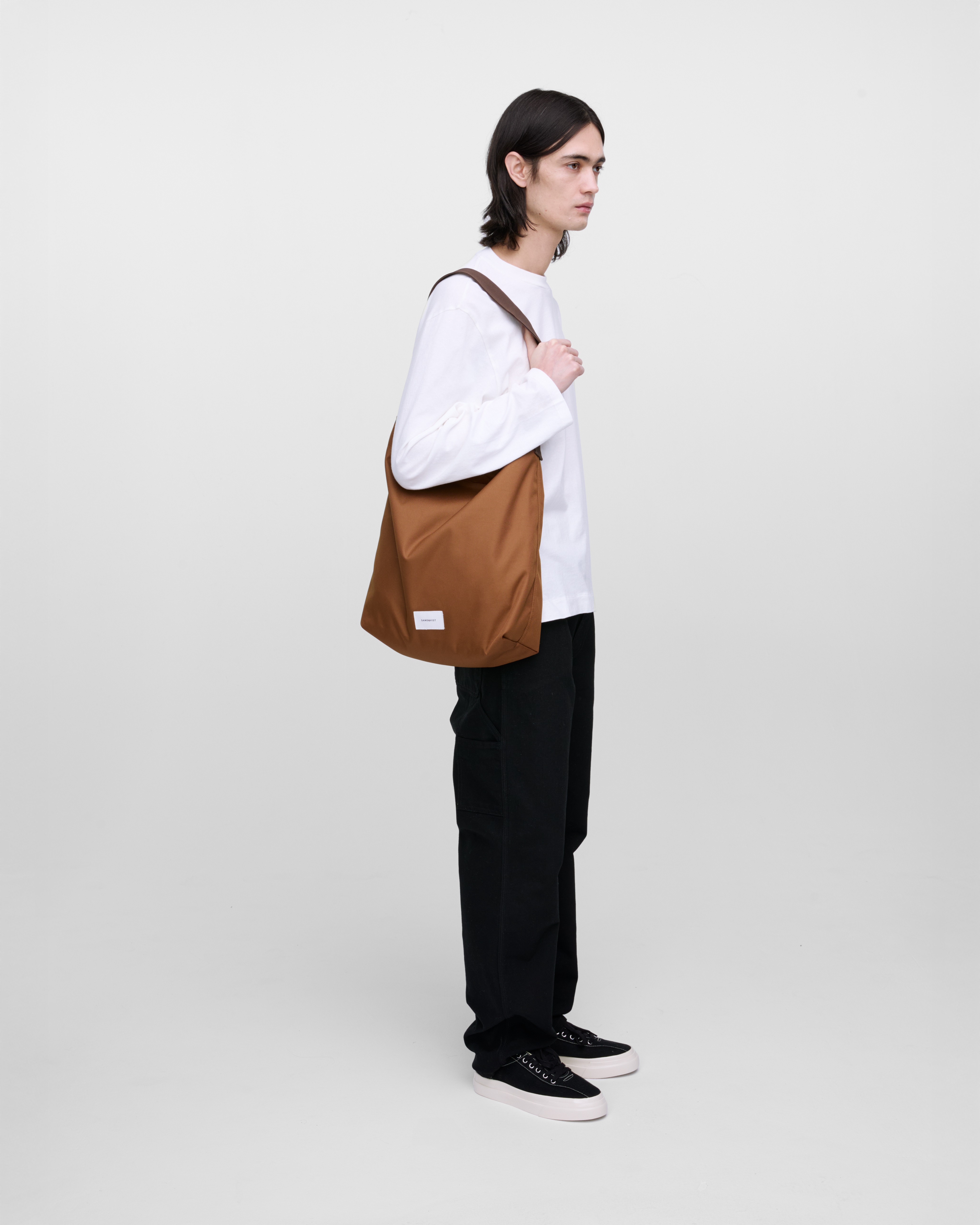 Mannequin masculin portant un sac Sandqvist Ground Shopper Bag, marron. Vue de profil, portant le sac en bandoulière sur une épaule.
