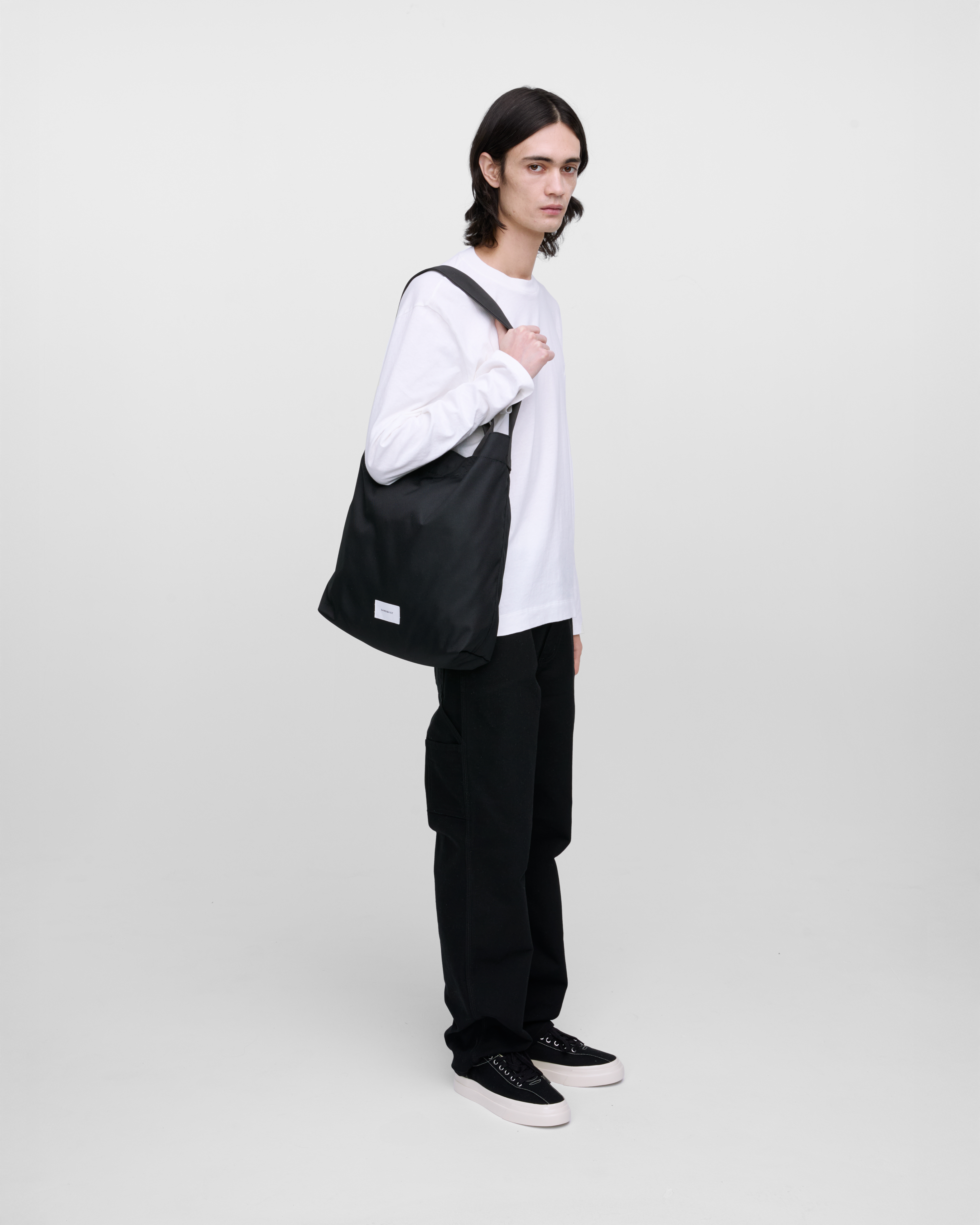 Male model wearing Sandqvist Ground Shopper Bag, black. Side view, wearing the bag on one shoulder.
