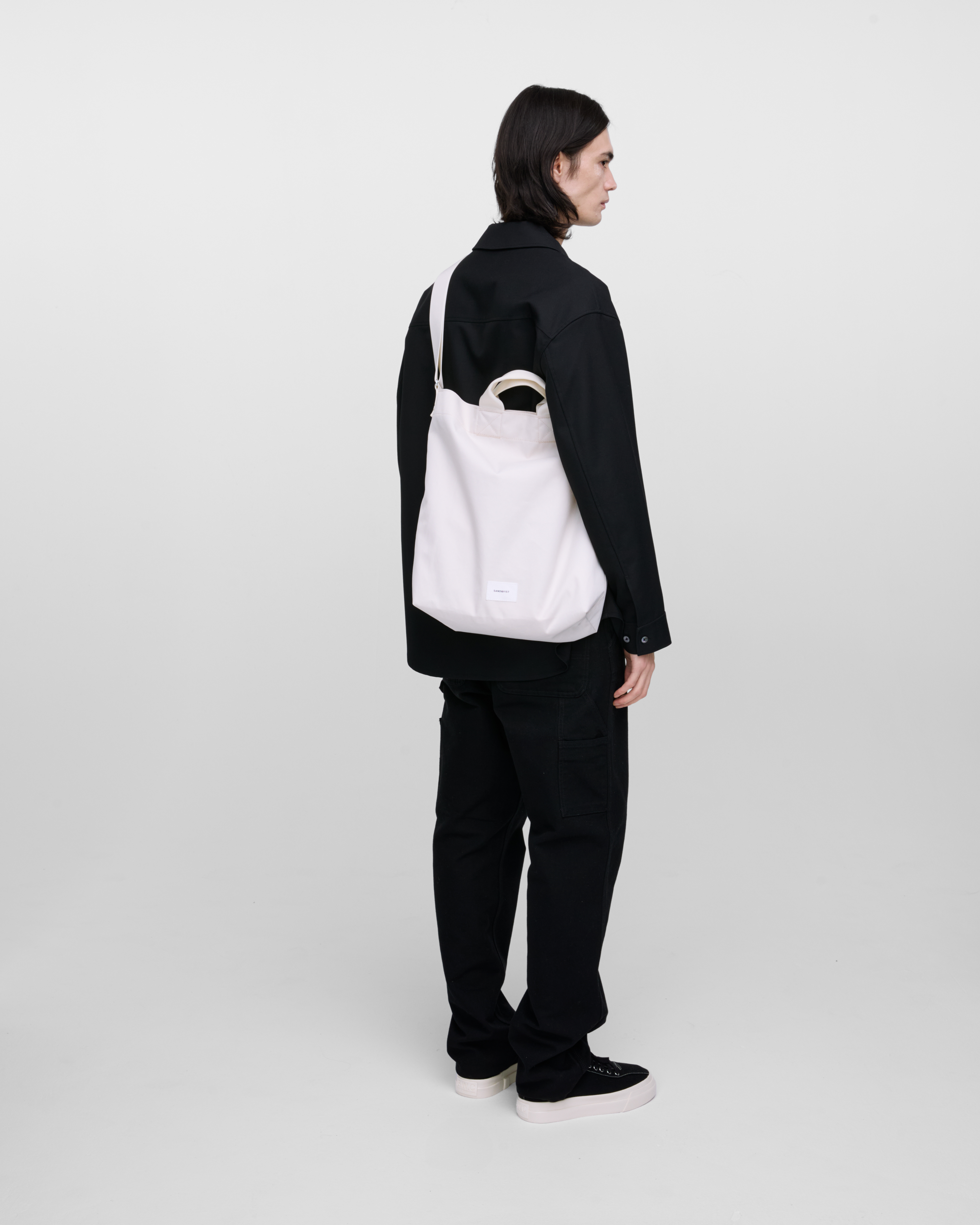 Male model wearing Sandqvist Ground Shopper Bag, white. Back view, wearing the bag on one shoulder crossbody.