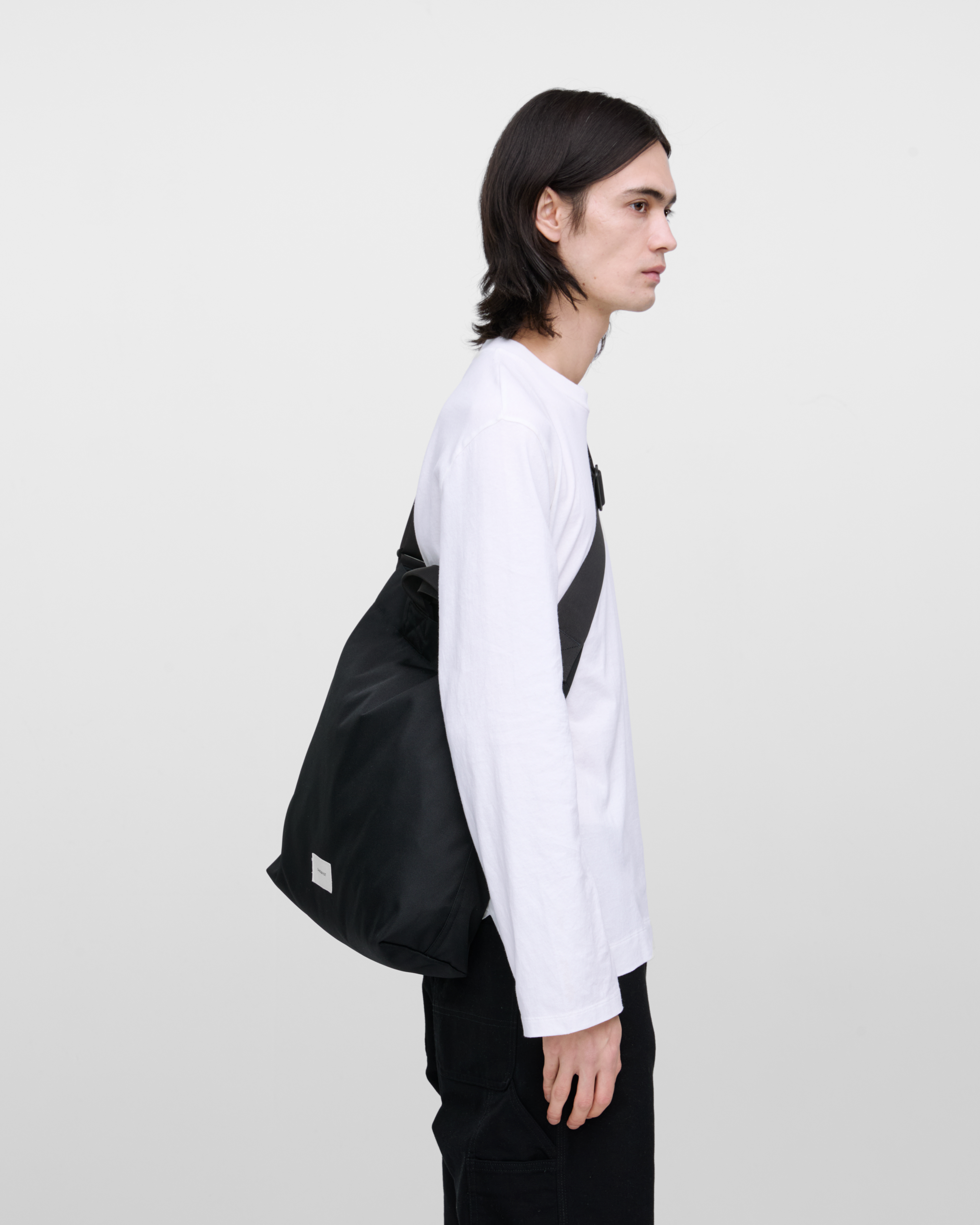 Male model wearing Sandqvist Ground Shopper Bag, black. Side view, wearing the bag on one shoulder crossbody.