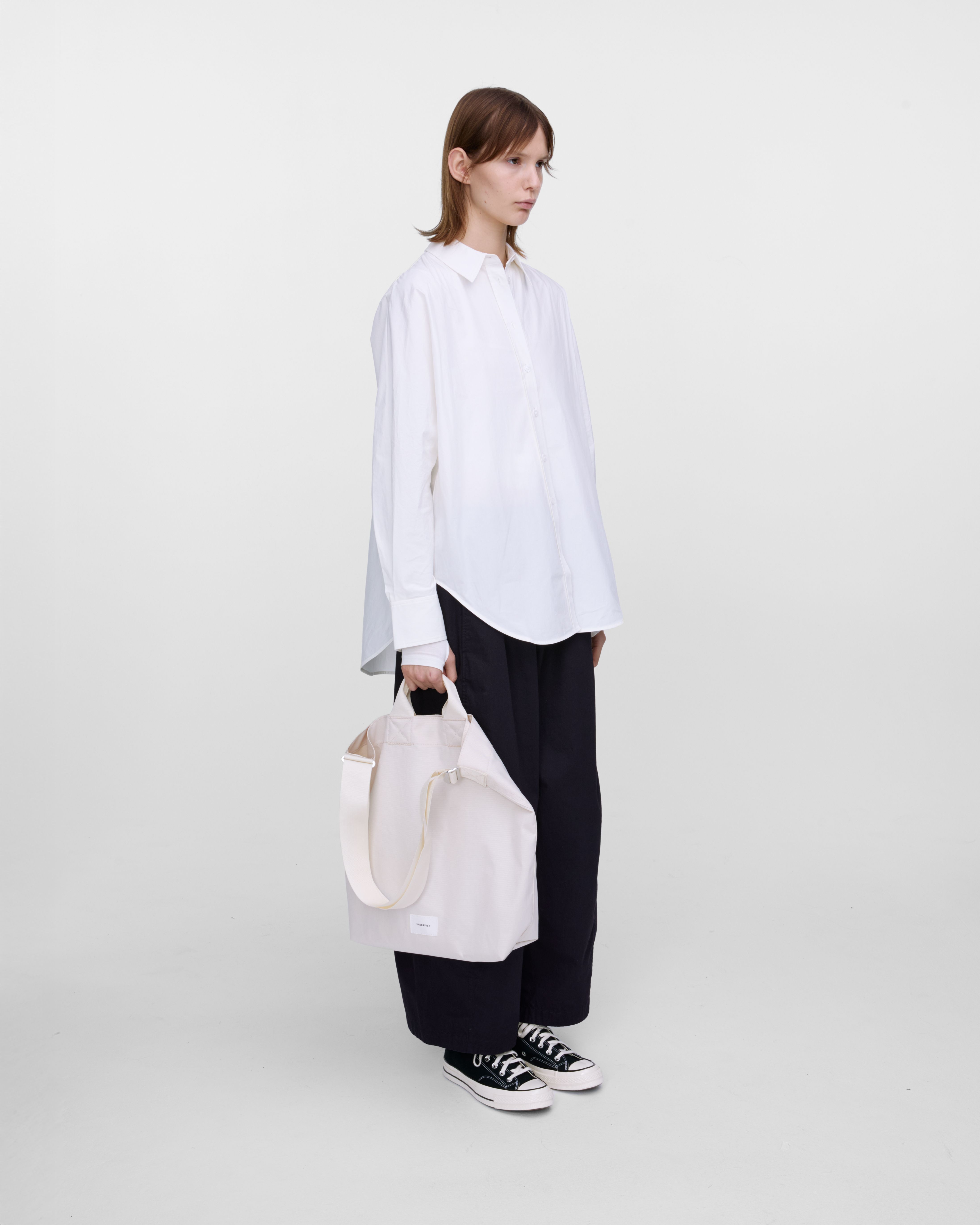 Female model holding Sandqvist Ground Shopper Bag, white. Side view, holding the bag in one hand.