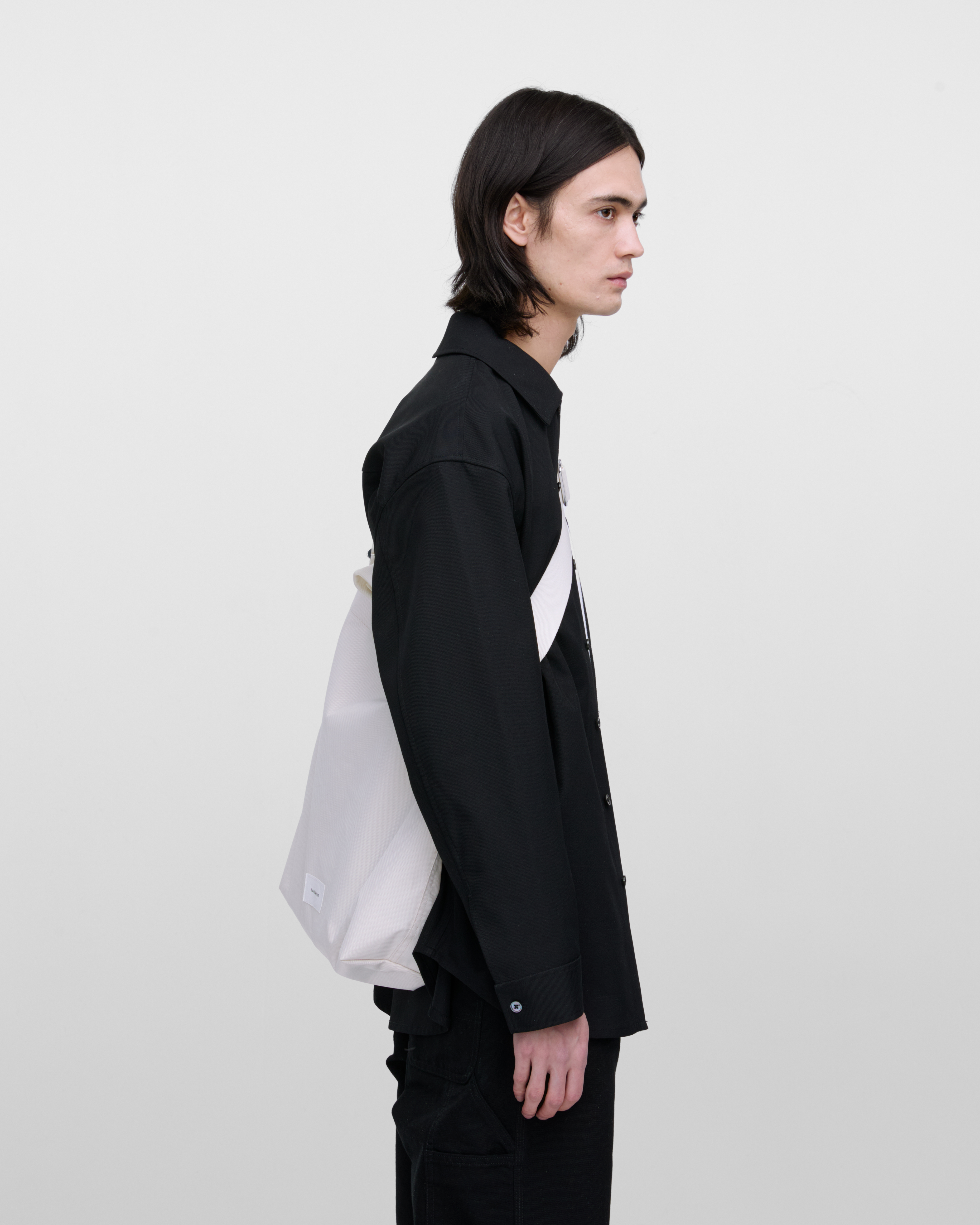 Male model wearing Sandqvist Ground Shopper Bag, white. Side view, wearing the bag on one shoulder crossbody.