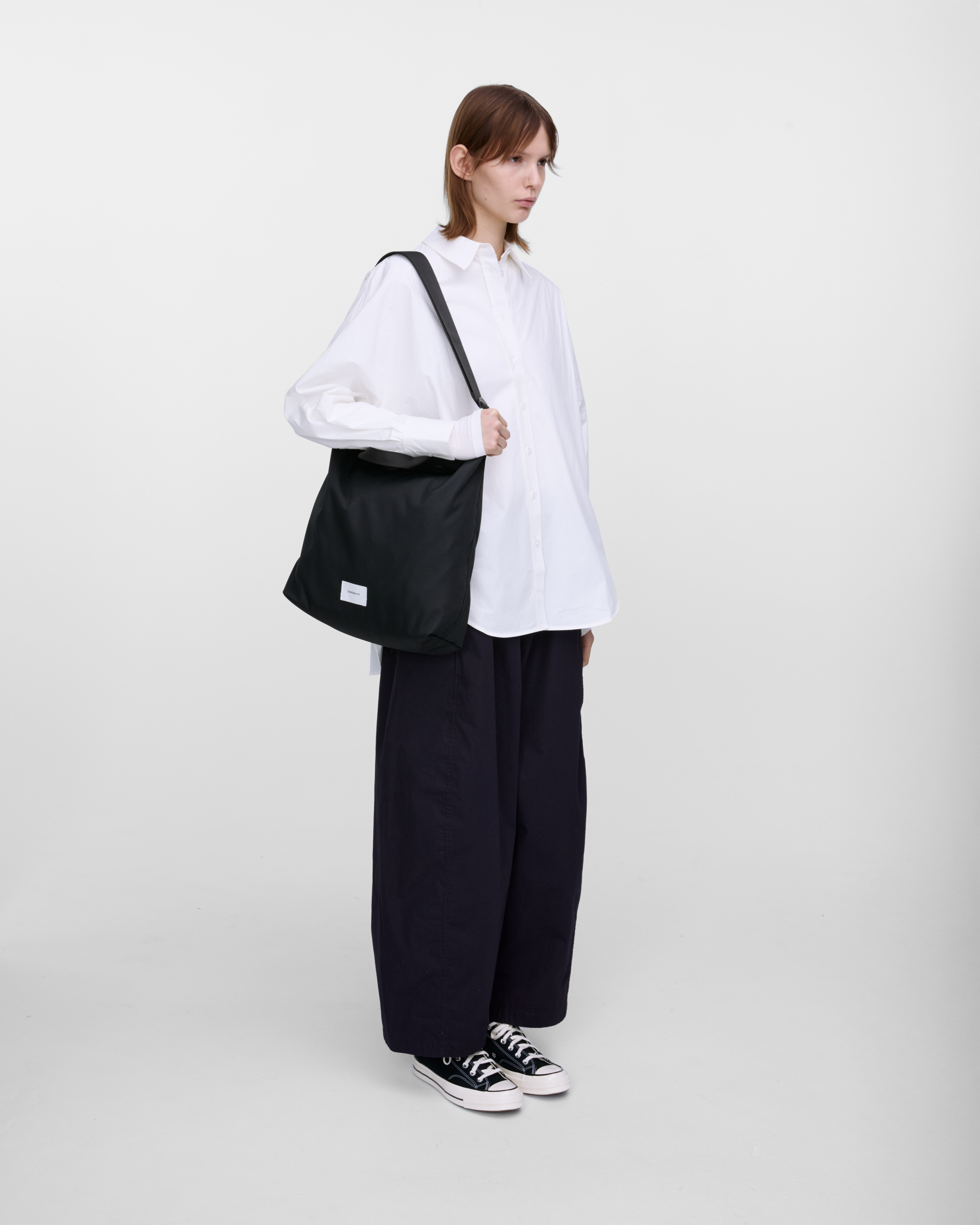 Female model wearing Sandqvist Ground Shopper Bag, black. Side view, wearing the bag on one shoulder.