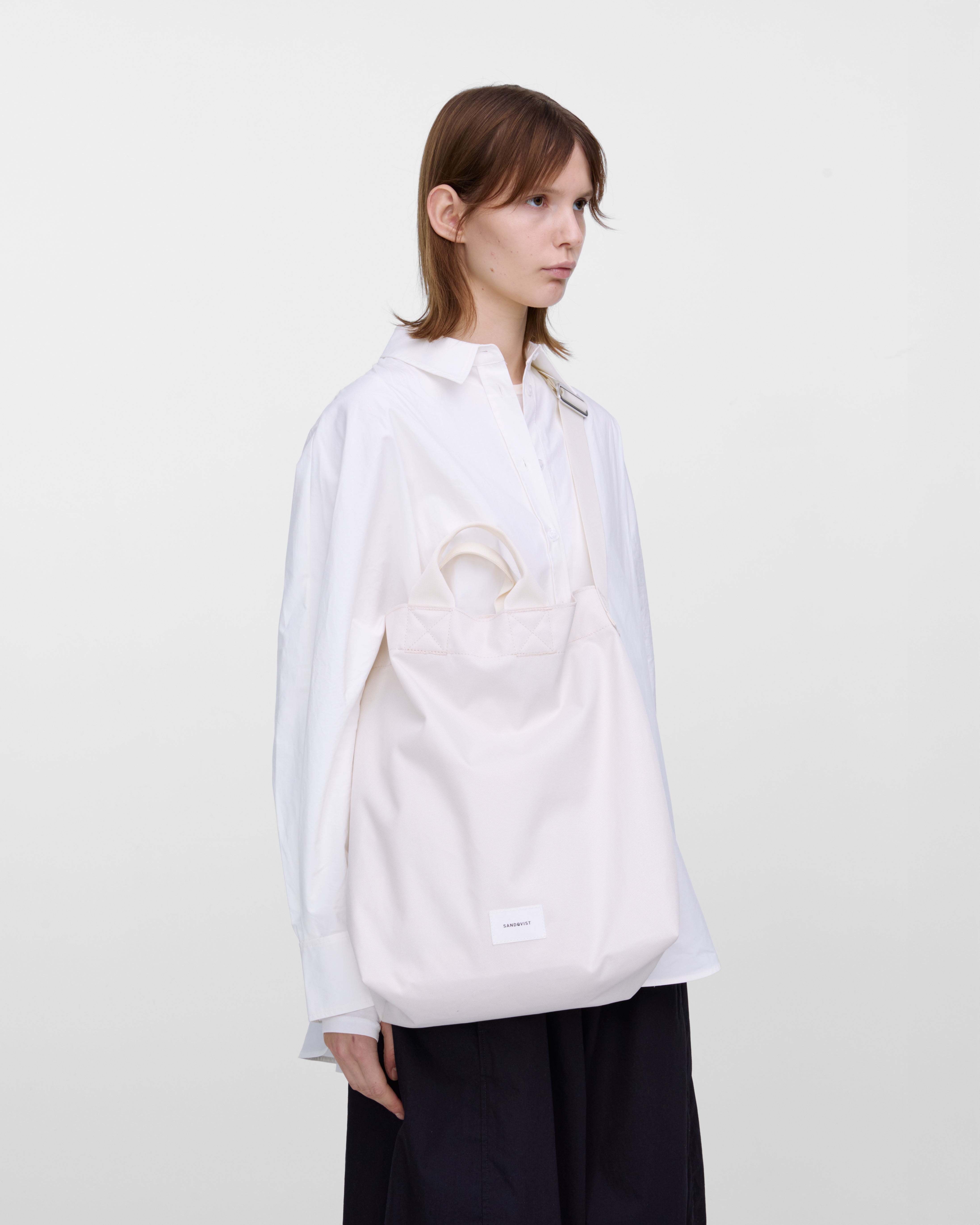 Female model wearing Sandqvist Ground Shopper Bag, white. Side view, wearing the bag on one shoulder crossbody.